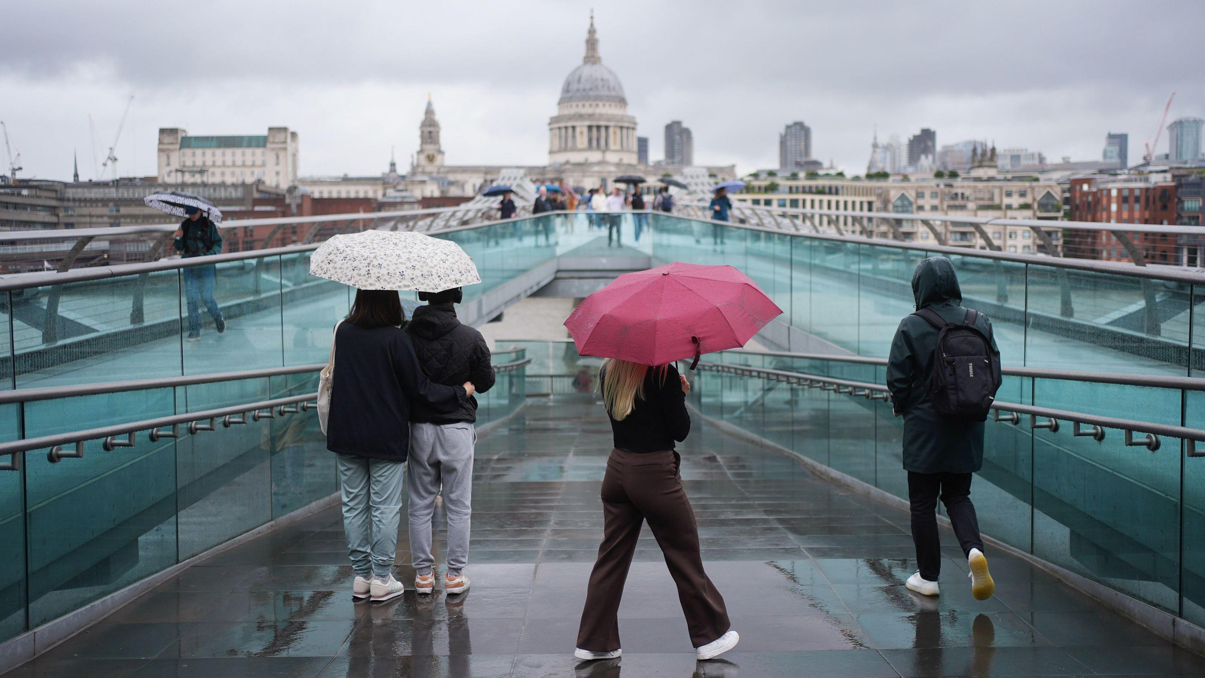 People with umbrellas walk over a bridge in the rain with the London skyline in the background