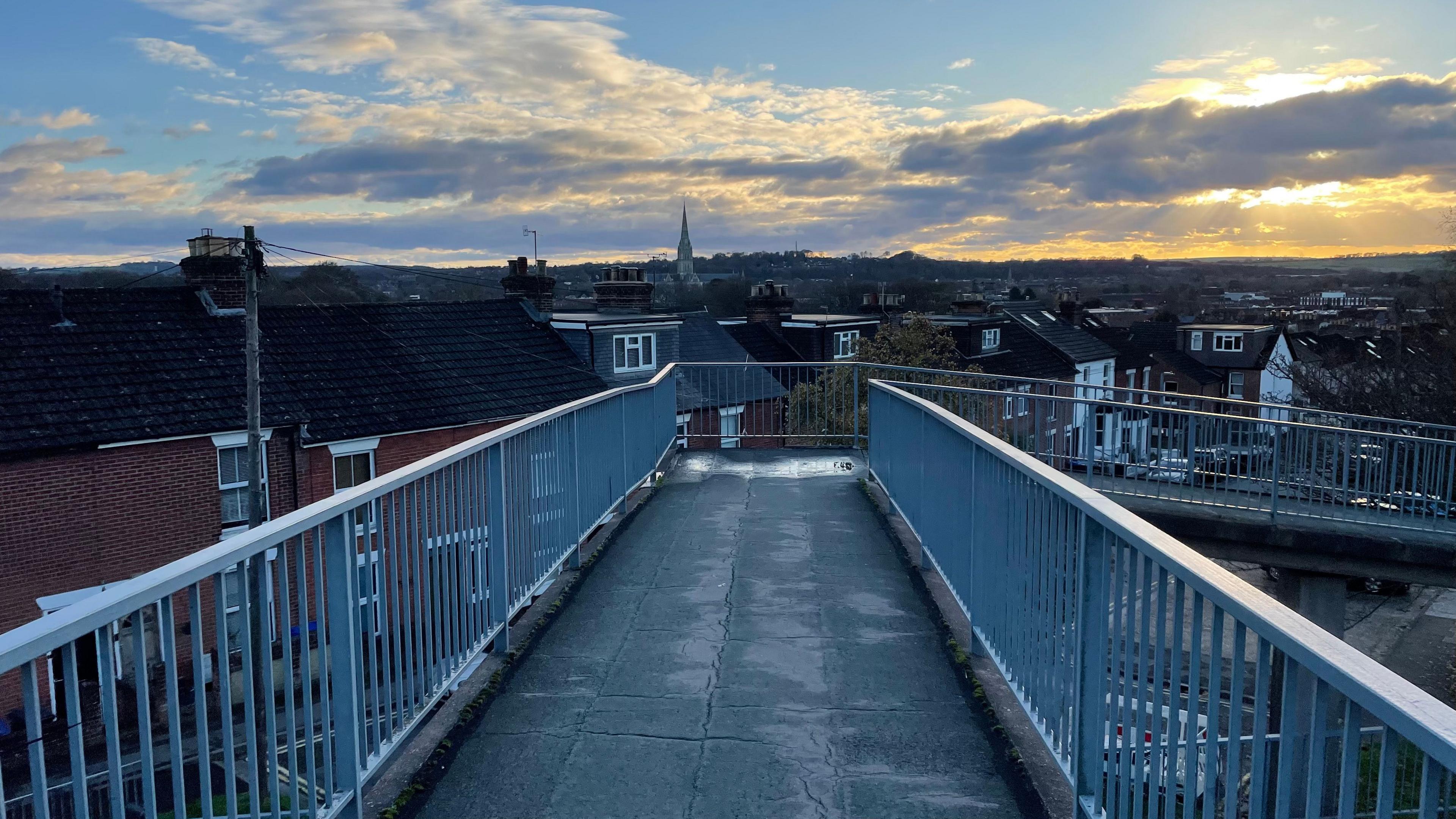 Footbridge POV shot, it is a concrete pathway with solid grey railings on either side and the skyscape of Salisbury in the background.