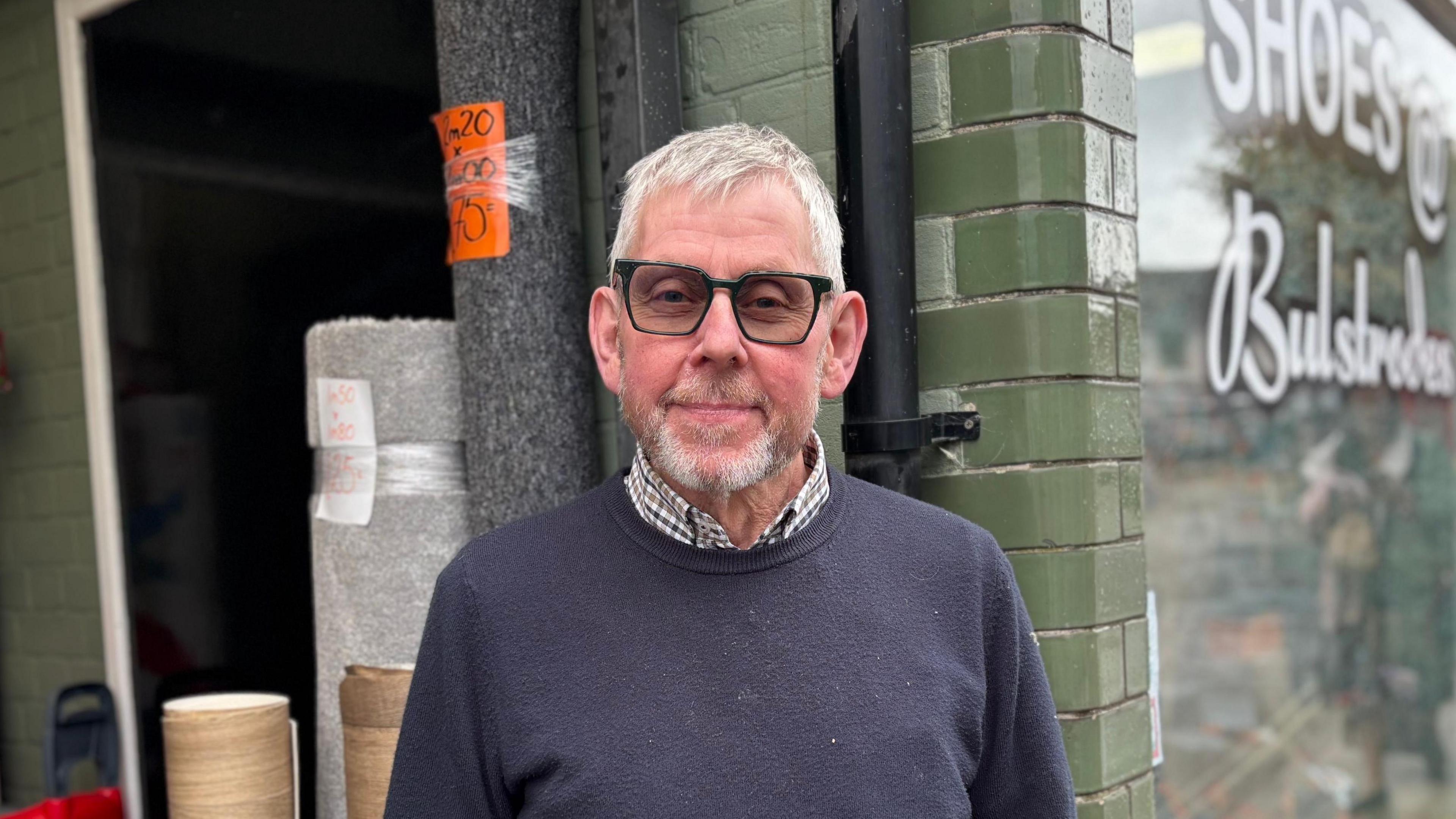A head and shoulders image of Bill Bulstrode, who is standing in front of rolls of carpet and a green wall. He is wearing a navy jumper over a shirt, as well as glasses. He is looking directly into the camera.