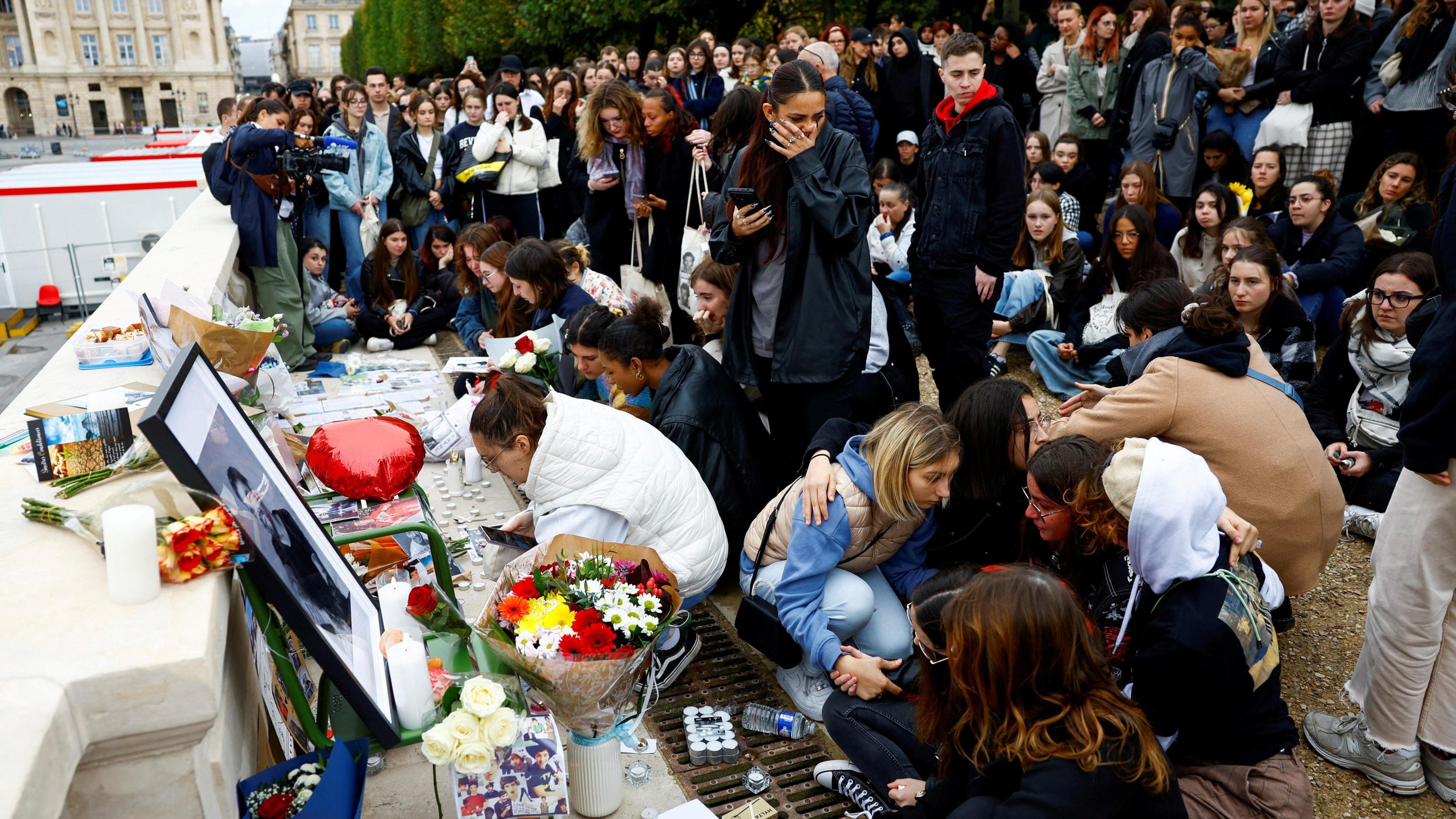 A crowd gathers in front of a framed photo of Liam Payne. Flowers and candles have been placed beneath. Some people are sitting on the floor in front of it and some fans are hugging each other.