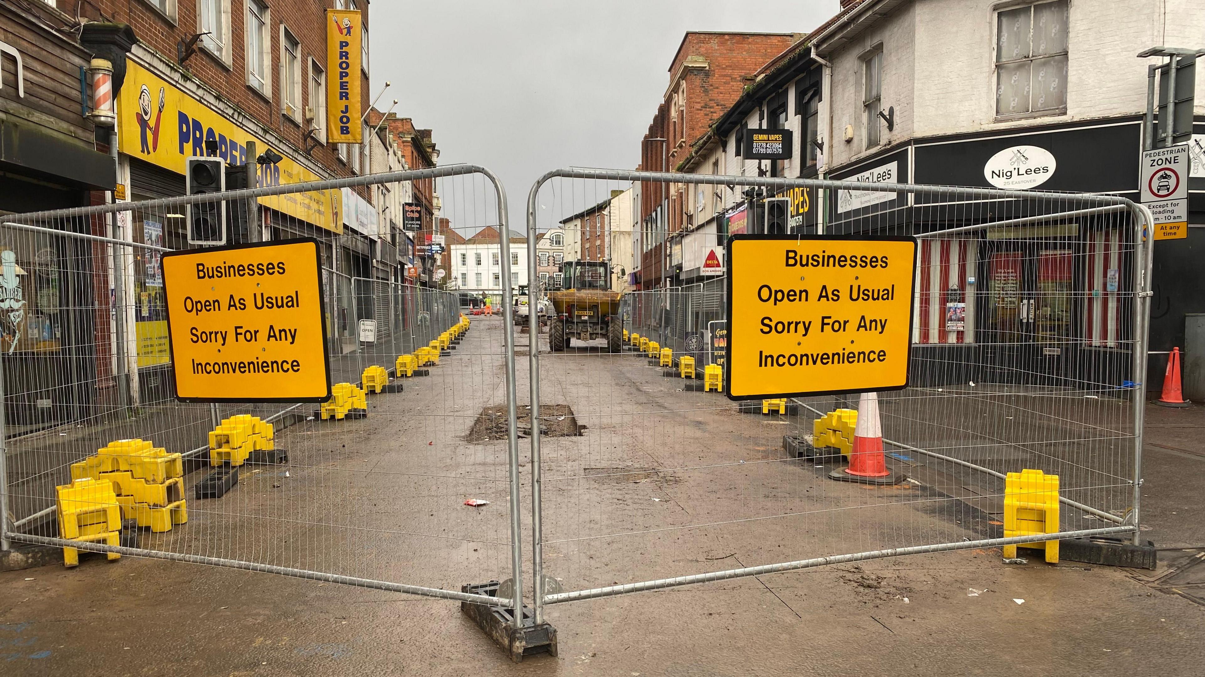 Fences barracade part of a street. Either side is various shops and inside the fencing is a tractor. On the fence signs it says 'businesses open as usual - sorry for any inconvenience' 