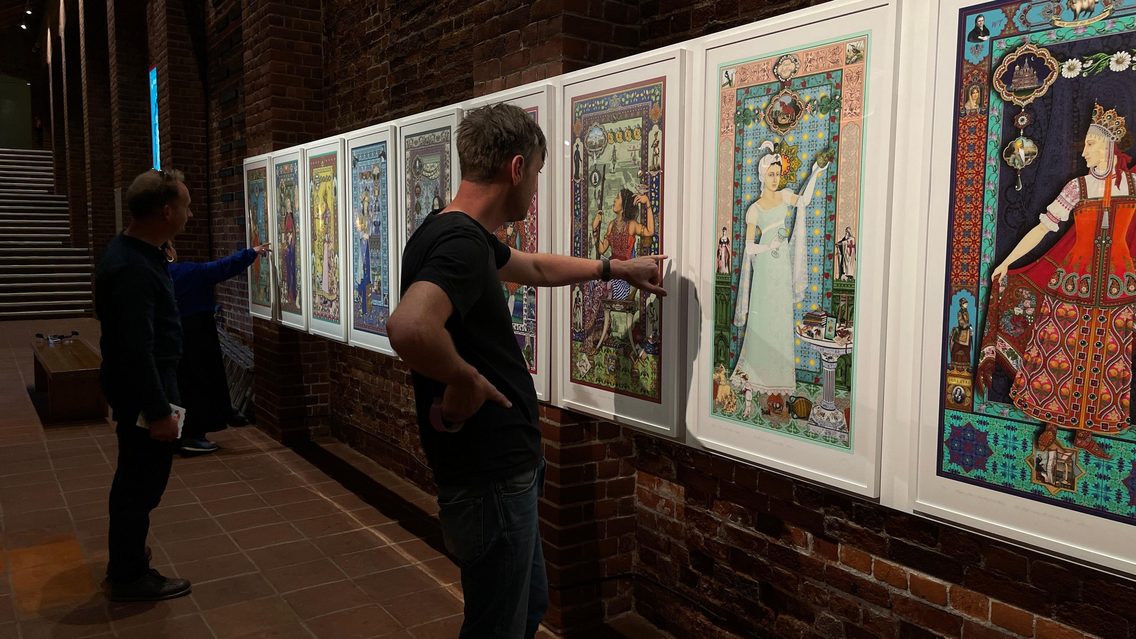 Two people stand and point at a row of white framed pictures. The pictures feature women in a series of highly patterned and brightly coloured works.
