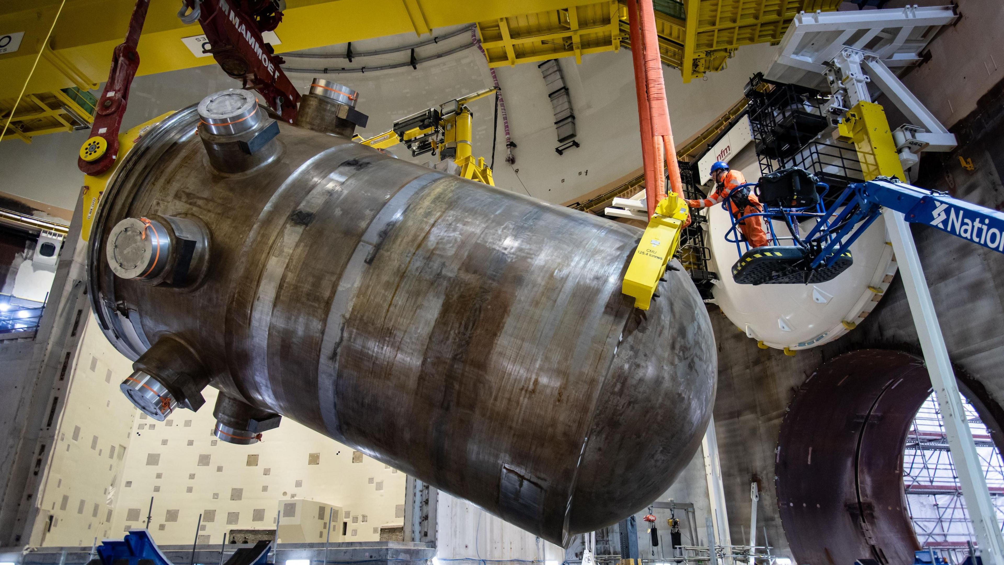 A giant grey steel cylinder hangs, half rotated in mid-air. Orange stays from an overhead crane hold it at both ends. A worker in high vis inspects it, he is tiny in comparison.