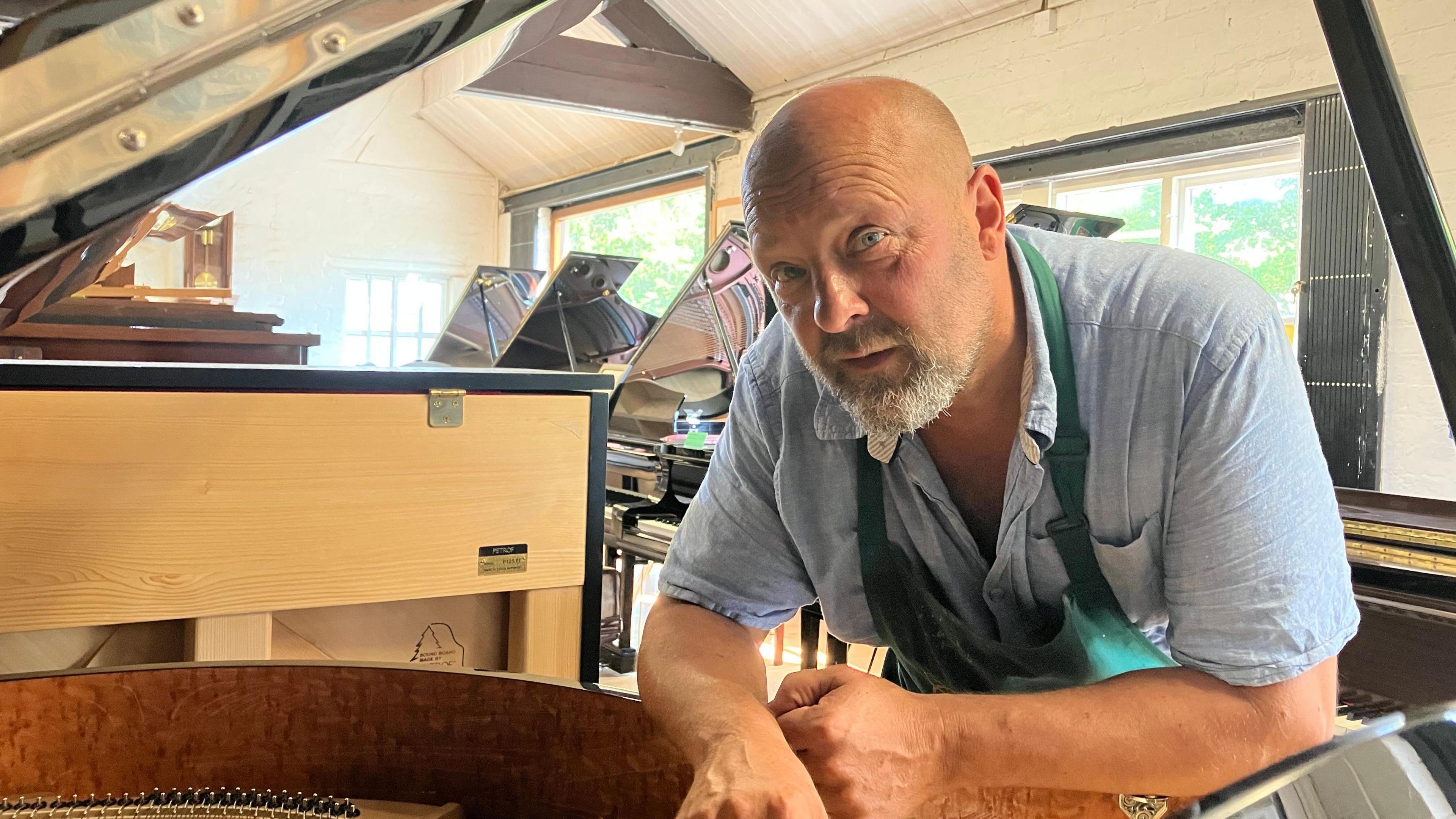 Adam Cox, owner of Yorkshire Pianos leans over a Cavendish piano in his showroom. He wears a blue short-sleeved shirt and a green apron. In the background, various pianos can be seen.