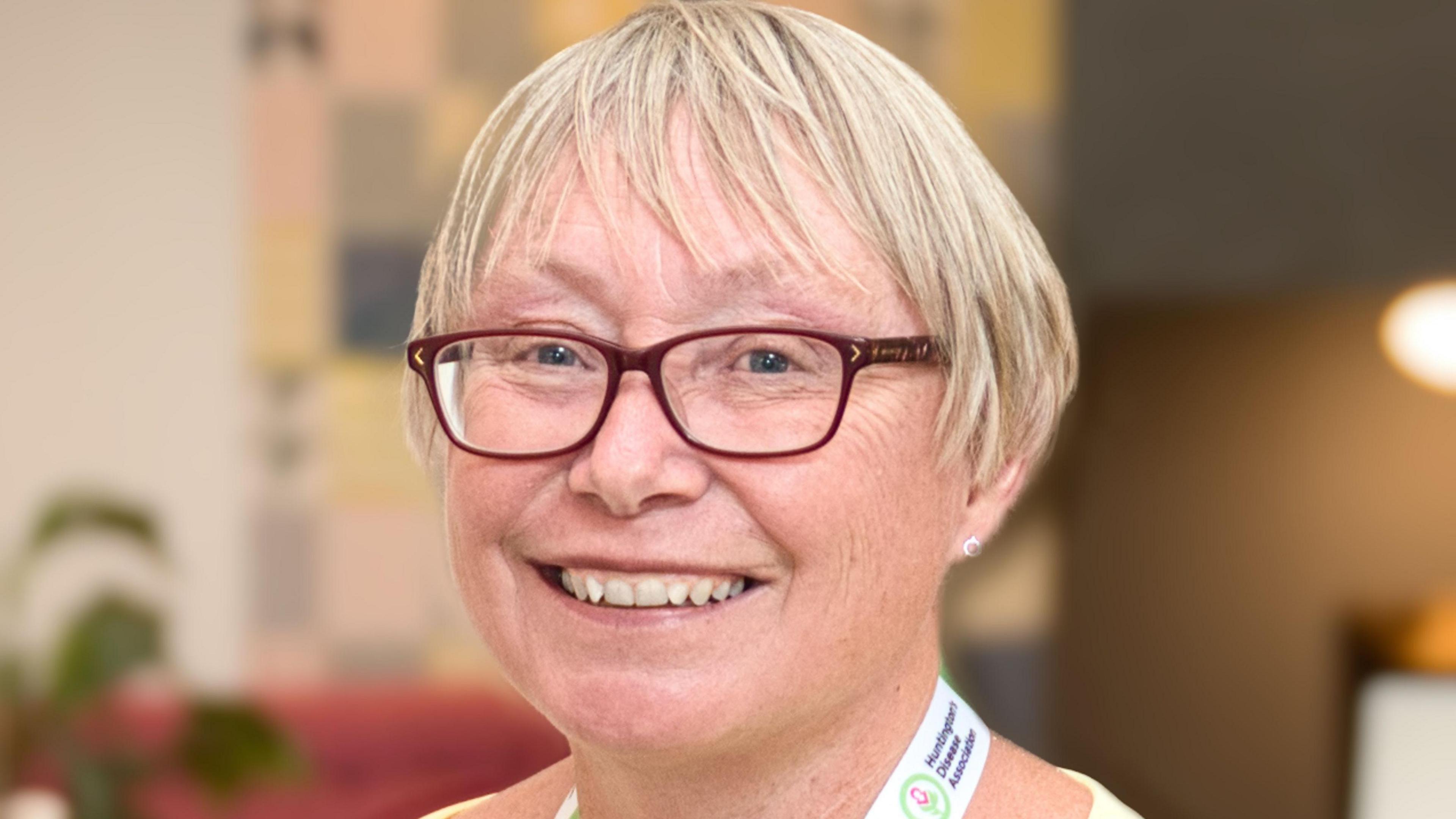 Cath is wearing glasses and a yellow top smiles at the camera. She has a lanyard around her neck with “Huntington’s Association” printed on it, and the background shows a warmly lit indoor setting