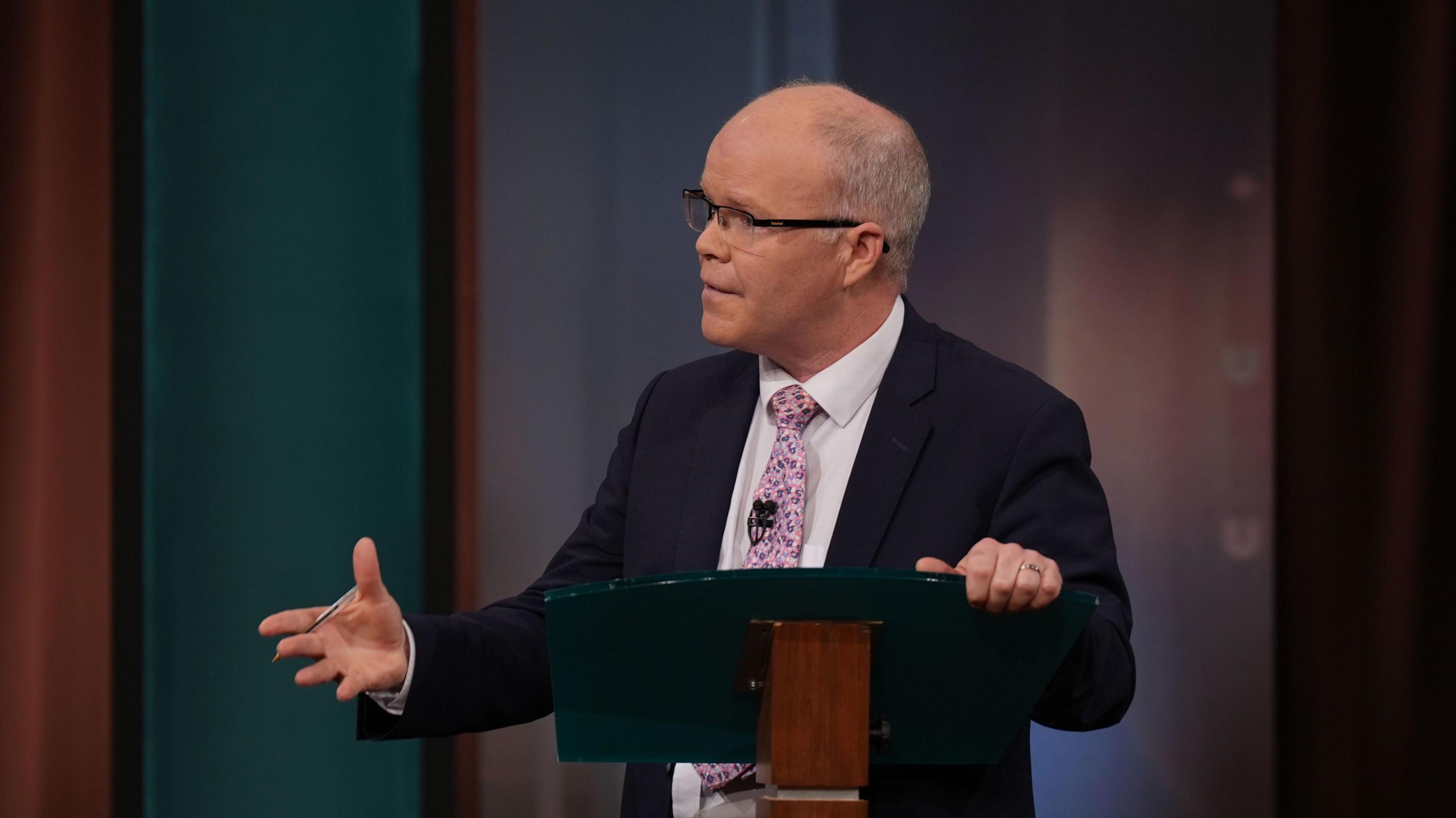 Leader of Aontu, Peadar Toibin during the General Election leaders' debate at RTE studios in Montrose, Dublin. He is standing at a podium. He is wearing glasses, wearing a navy blazer, white shirt and pink tie.