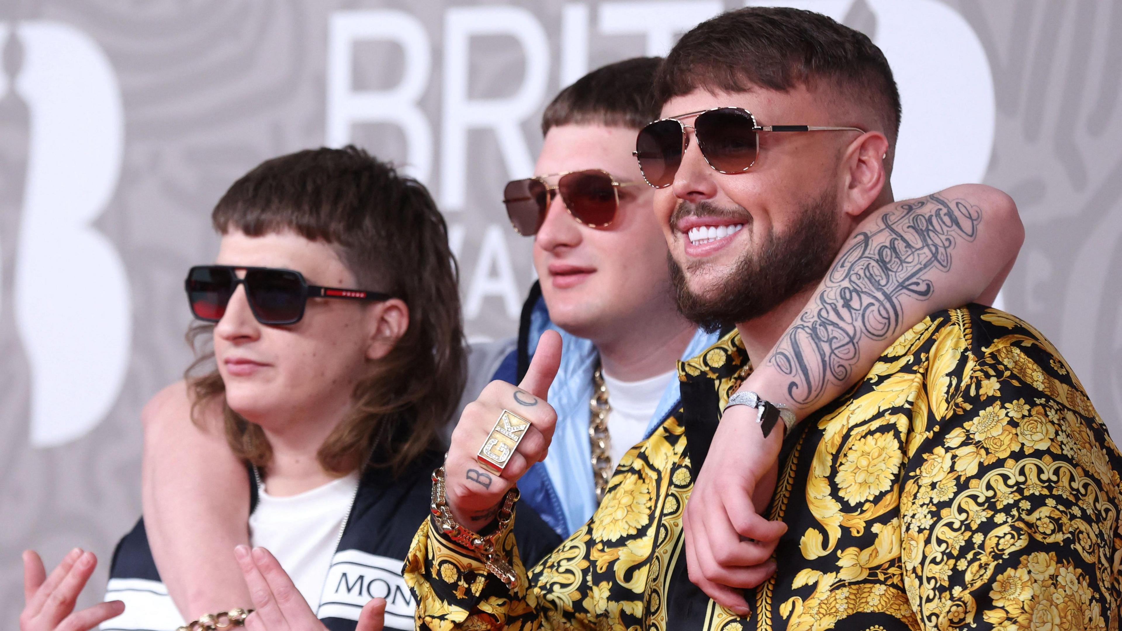 Bad Boy Chiller Crew are on the red carpet at the Brits in 2023. They are posing for photographers, wearing dark sunglasses. Clive is gesturing with his hands. Kane has his arms draped around the other men's necks as they stand either side of him. GK is smiling with his thumb up and wearing a heavily-patterned black and gold jacket.