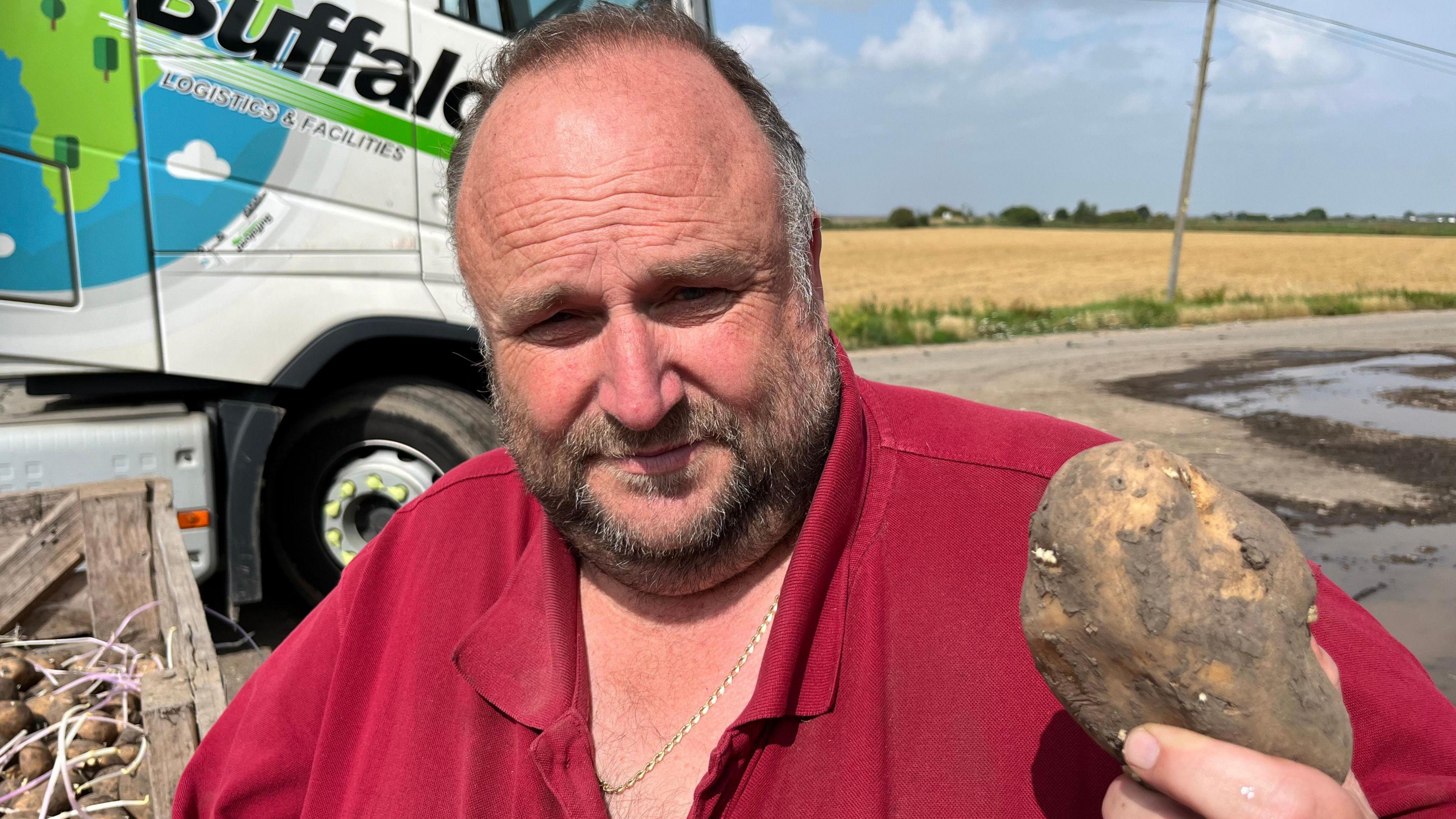 Mr Parson is holding a potato up to the camera. He is wearing a red top and a gold chain around his neck. He has dark hair and facial hair. Behind him is a field and potatoes in crates. They have grown sprouts due to being in storage for so long.
