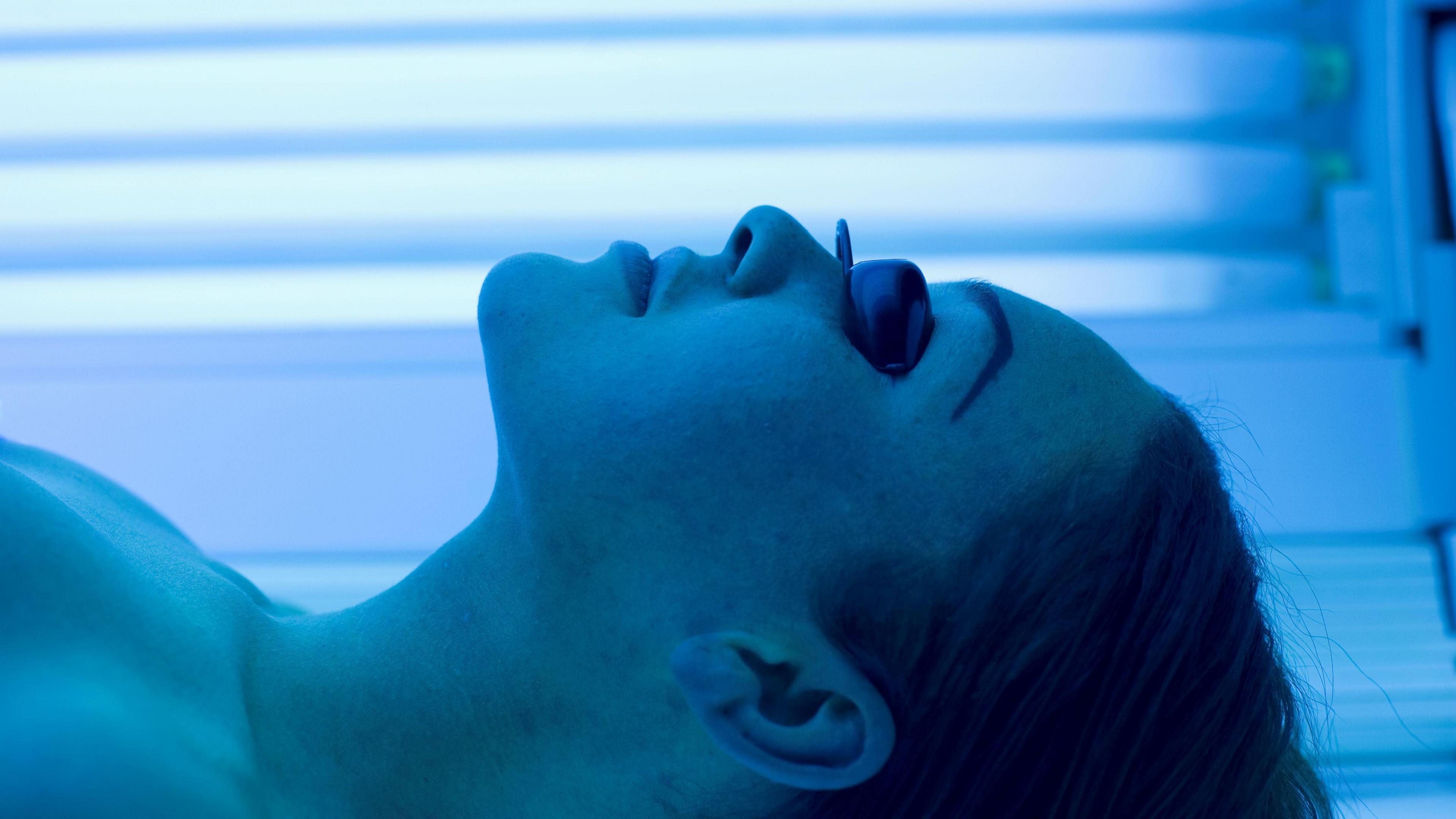 A woman is wearing goggles to protect her eyes. She's lying on a lie down sunbed. Blue lamps are shining on her.