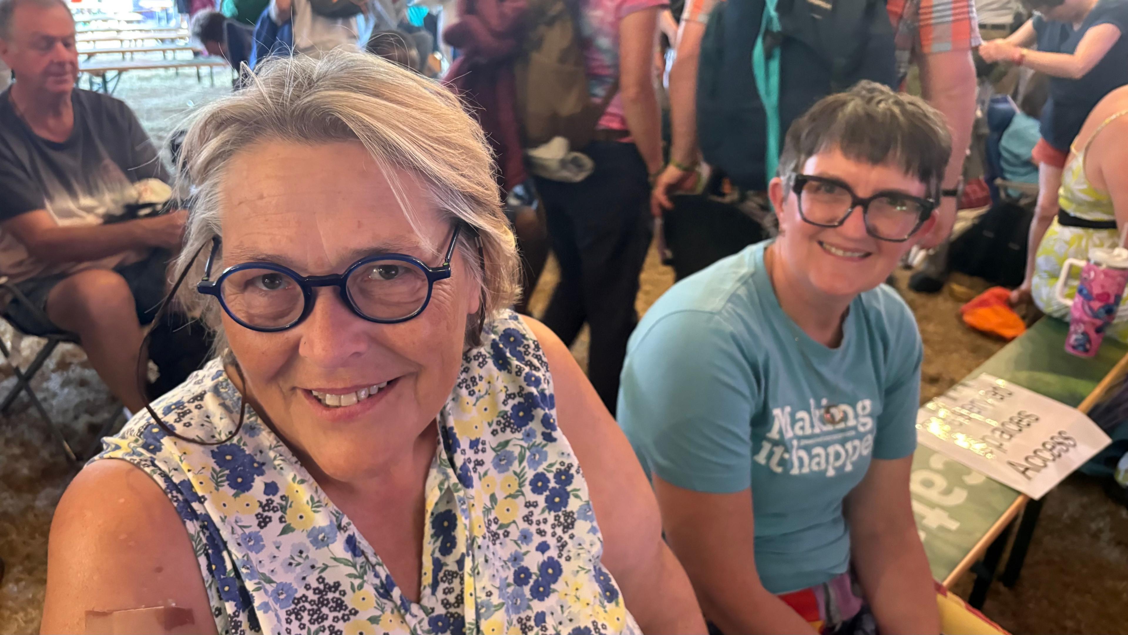Rachel with medium-length blonde hair and black glasses wearing a blue and yellow dress and Gillian with short dark hair and large black-framed glasses wearing a blue Making it Happen T-shirt sitting on a bench. There are other people walking behind them