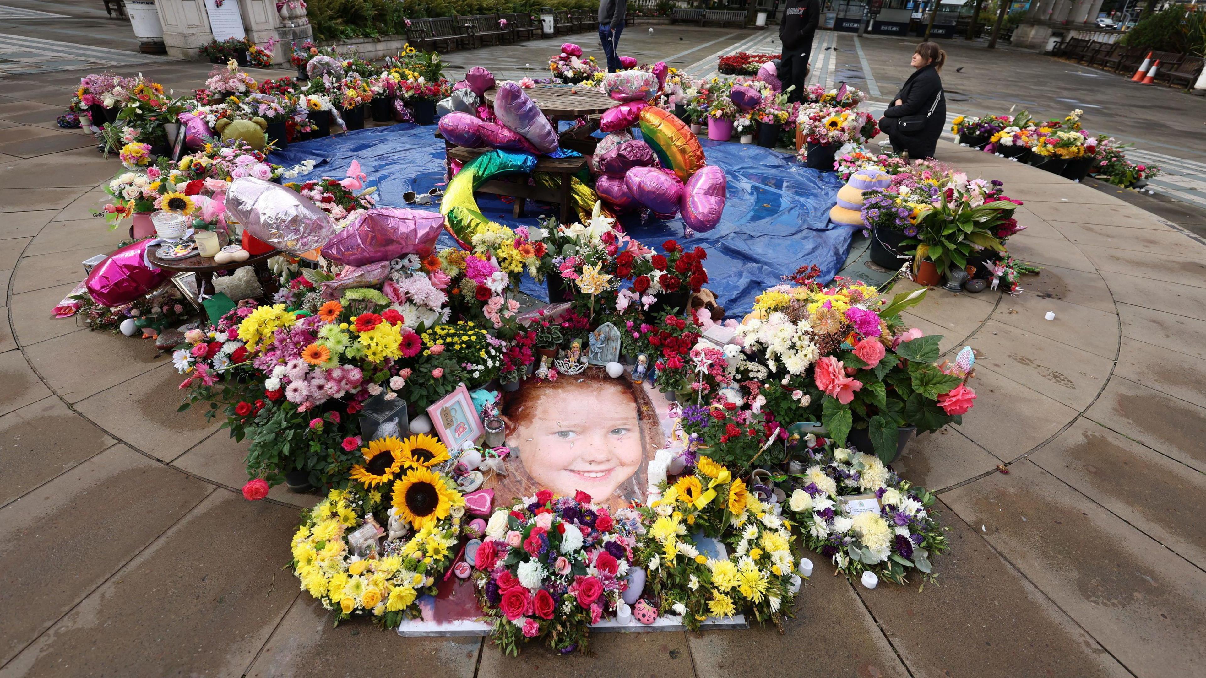 Dozens of bunches of flowers, colourful balloons and cards are laid out on a paved area, with a large picture of a smiling Elsie Dot Stancombe in front of the camera.