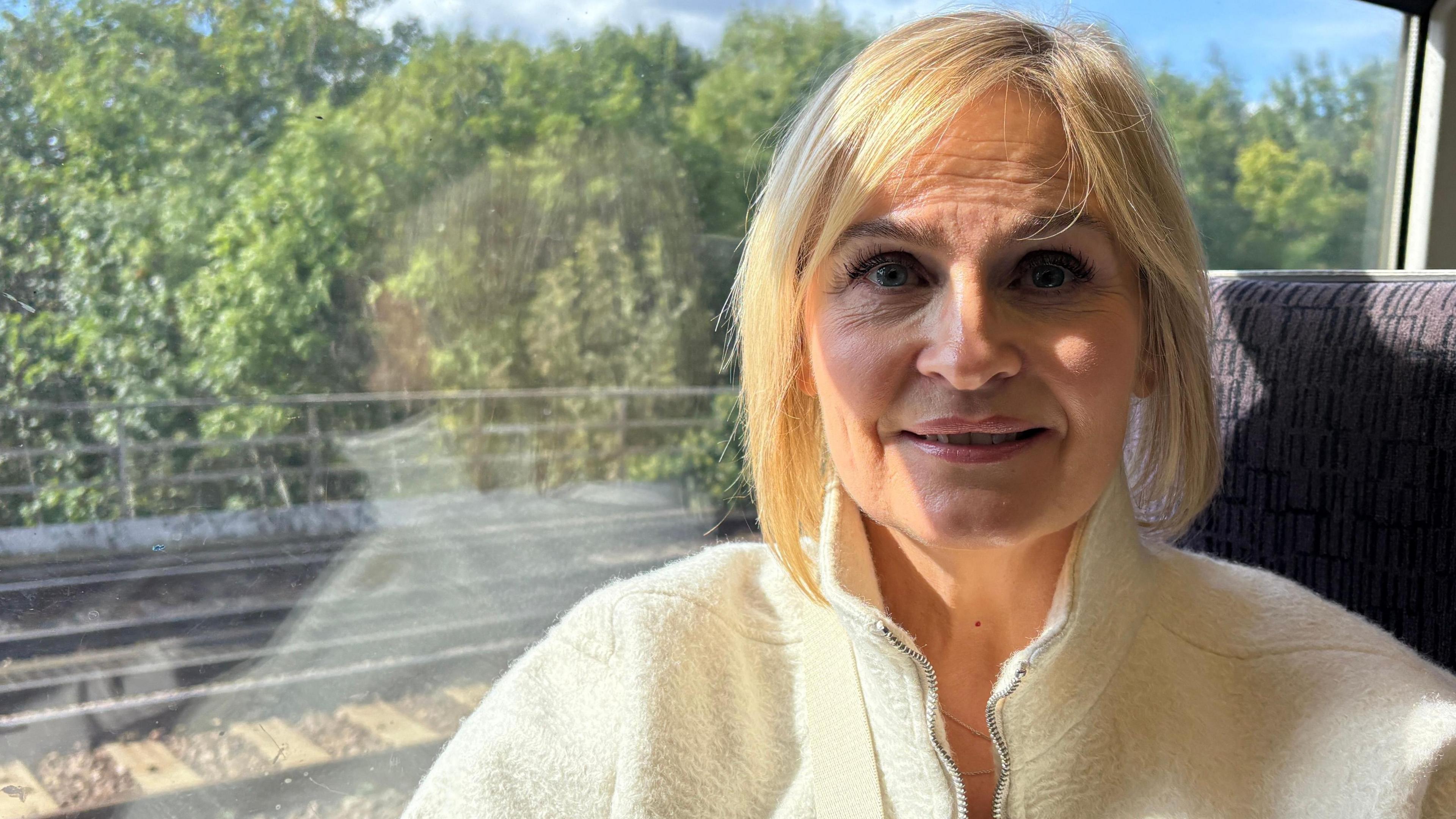 Natalie Barron looking directly down the camera. She is smiling slightly and is sitting next to a window inside a train carriage. She has short hair cut into a bob and is wearing a thick, white jacket. Part of a train line, trees and blue sky can be seen out the window next to her.