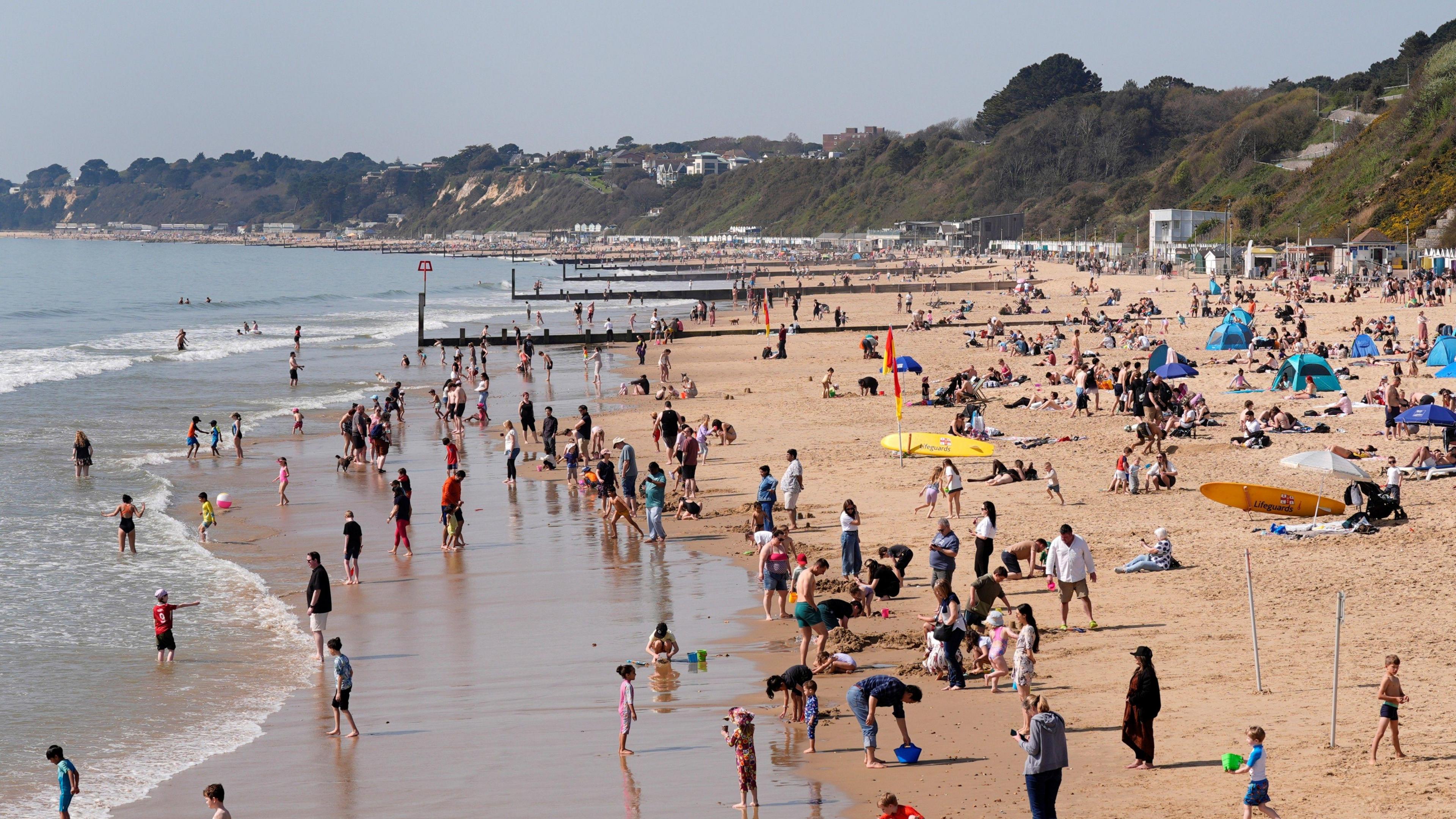 A photo of a busy beach with people on the sand and in the sea, with blue skies overhead.