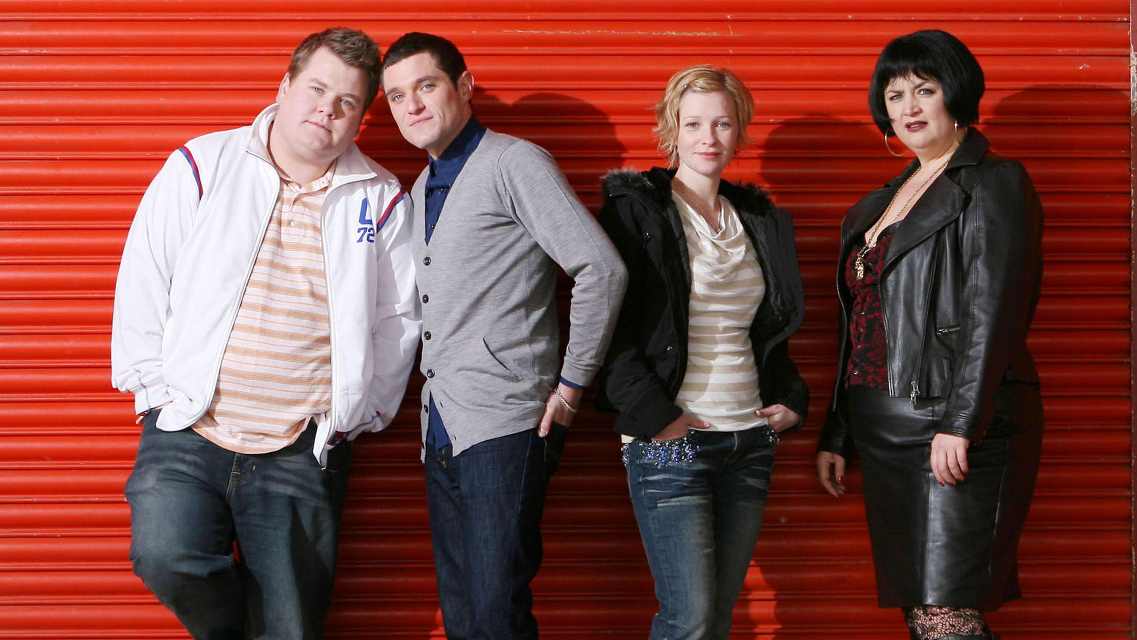 The cast of Gavin and Stacey assemble for an official cast photo ahead of the first ever episode. They are stood against a red shutter. Smithy (played by James Corden) is wearing a white jacket, over a white and mustard stiped t-shirt and blue jeans, he has his arms in his pockets and has one leg against the shutter and his leaning to his right, Gavin (played by Matthew Horne) is next to him, wearing a grey cardigan over a blue t-shirt with his left hand in the back pocket of his blue jeans. He is leaning into James Corden. Next to him is Stacey (played by Joanna Page), she is wearing a white and mustard t-shirt and a black coat, along with blue jeans and she has her hands in her pockets, she is stood next to Nessa (played by Ruth Jones), who is wearing a black leather jacket and a black leather shirt, with a red and black top underneath. 