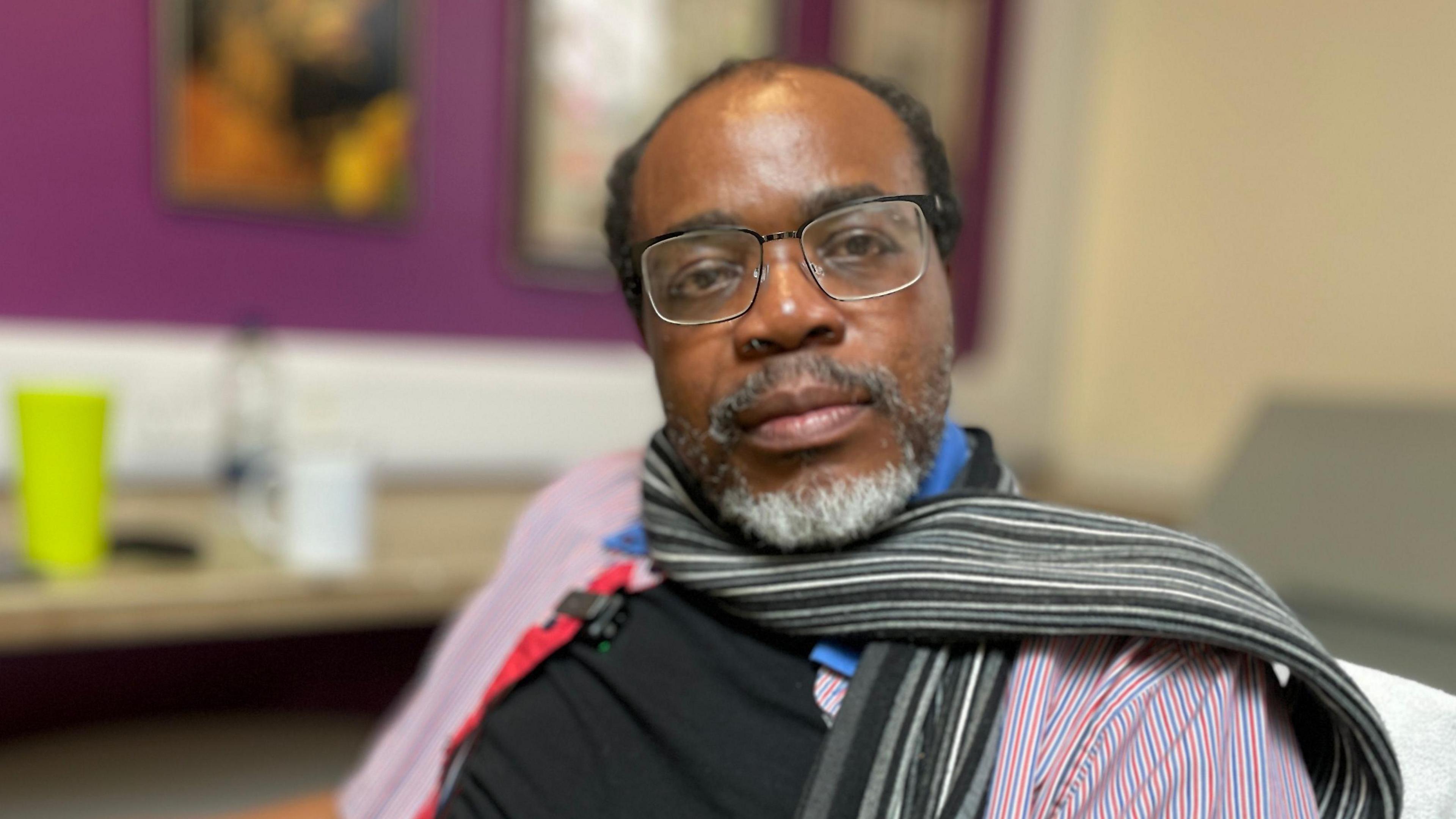 A man sitting down wearing black frame glasses. He is wearing a black scarf and a blue t-shirt with a shirt over the top. He is looking at the camera. He is not smiling but does not look unhappy either.