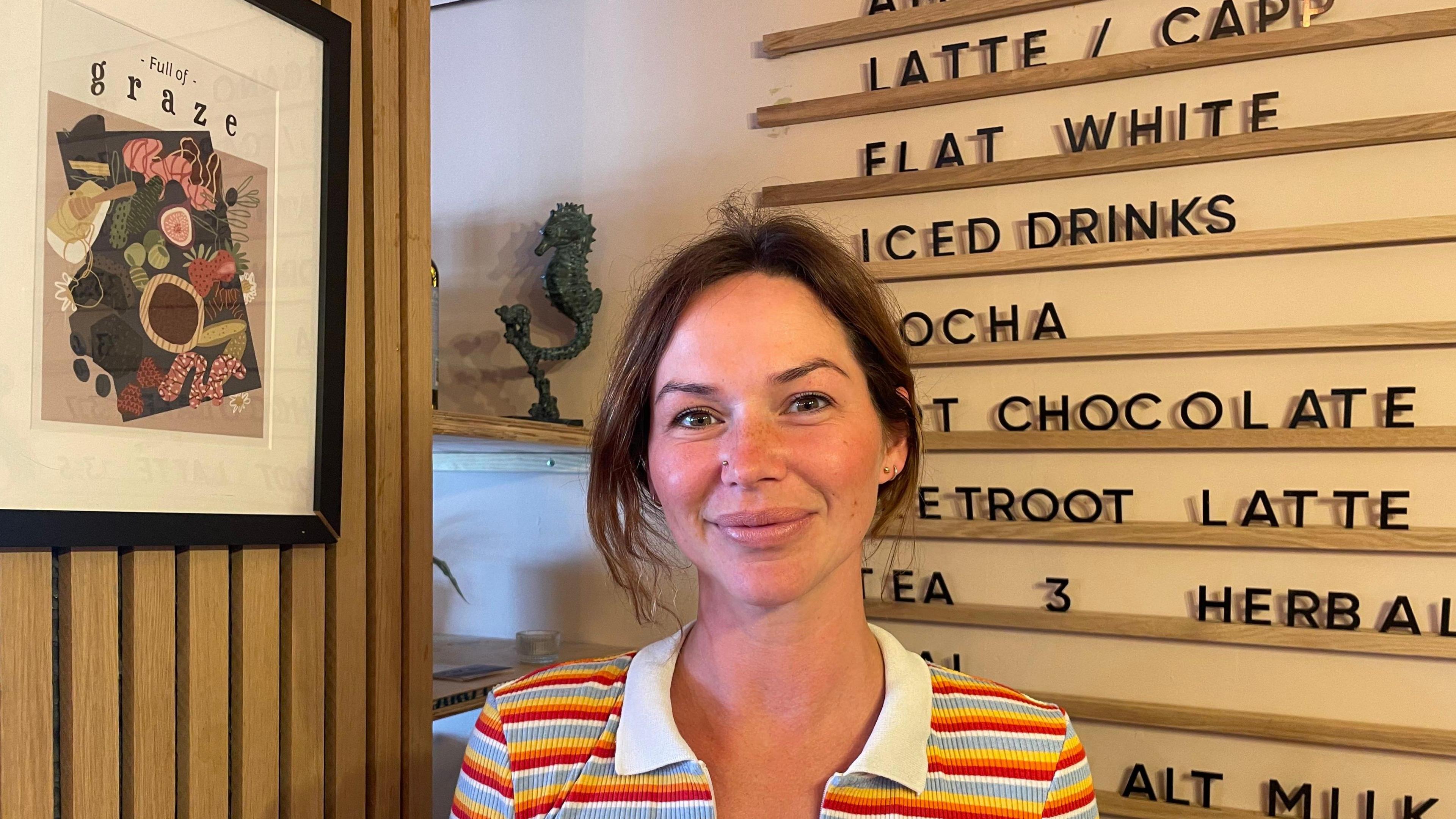 Jenny Morgan wearing a colourful stripy top and stood in front of a board which lists different types of drinks on sale