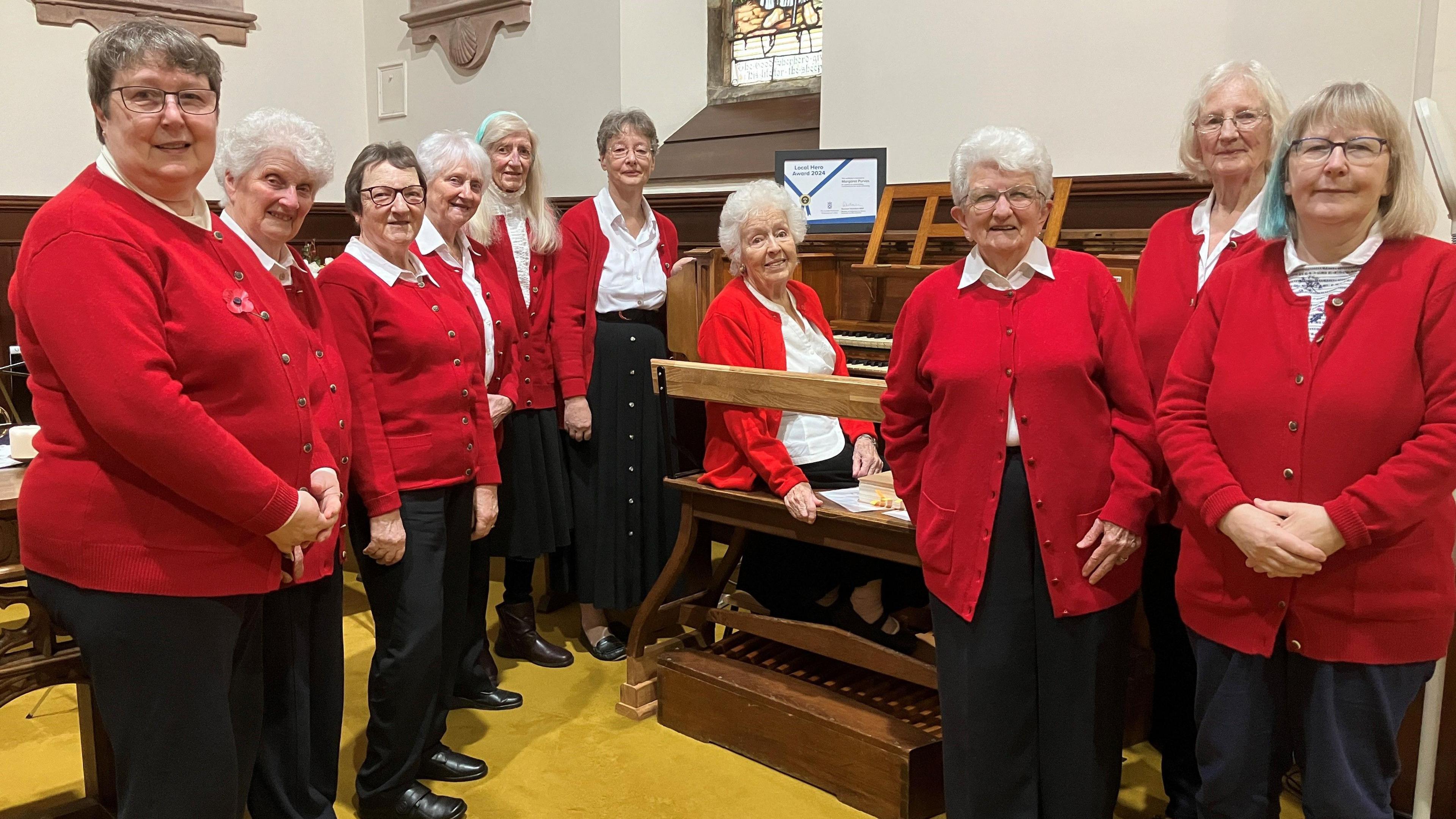 A choir of women in white blouses, red cardigans and black trousers and skirts stand around a white-haired woman sitting at a church organ