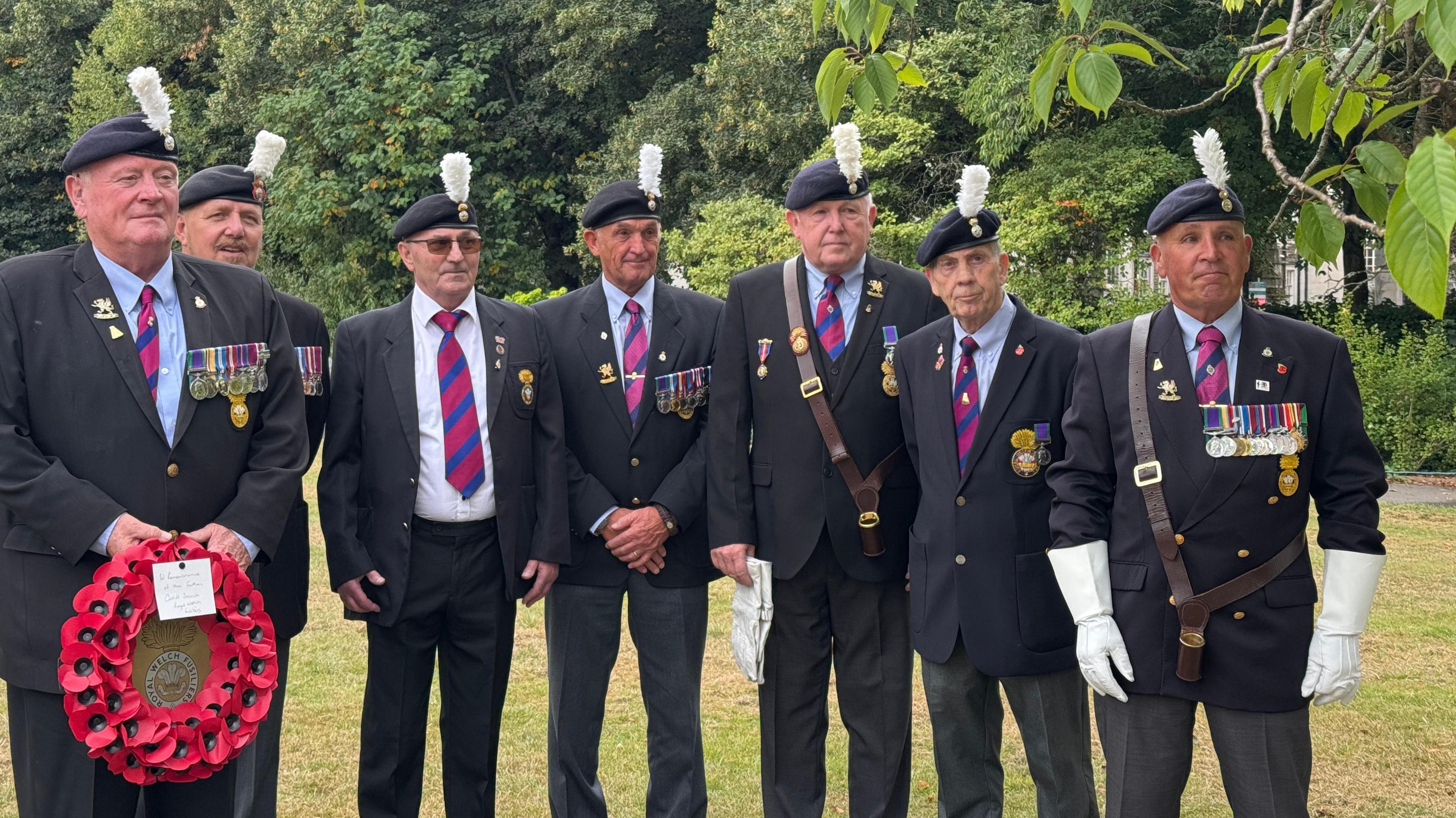 Photo of a group of servicemen in their full uniform, which includes a three piece suit and a red and navy stripe tie. They all stand in a line side by side. 