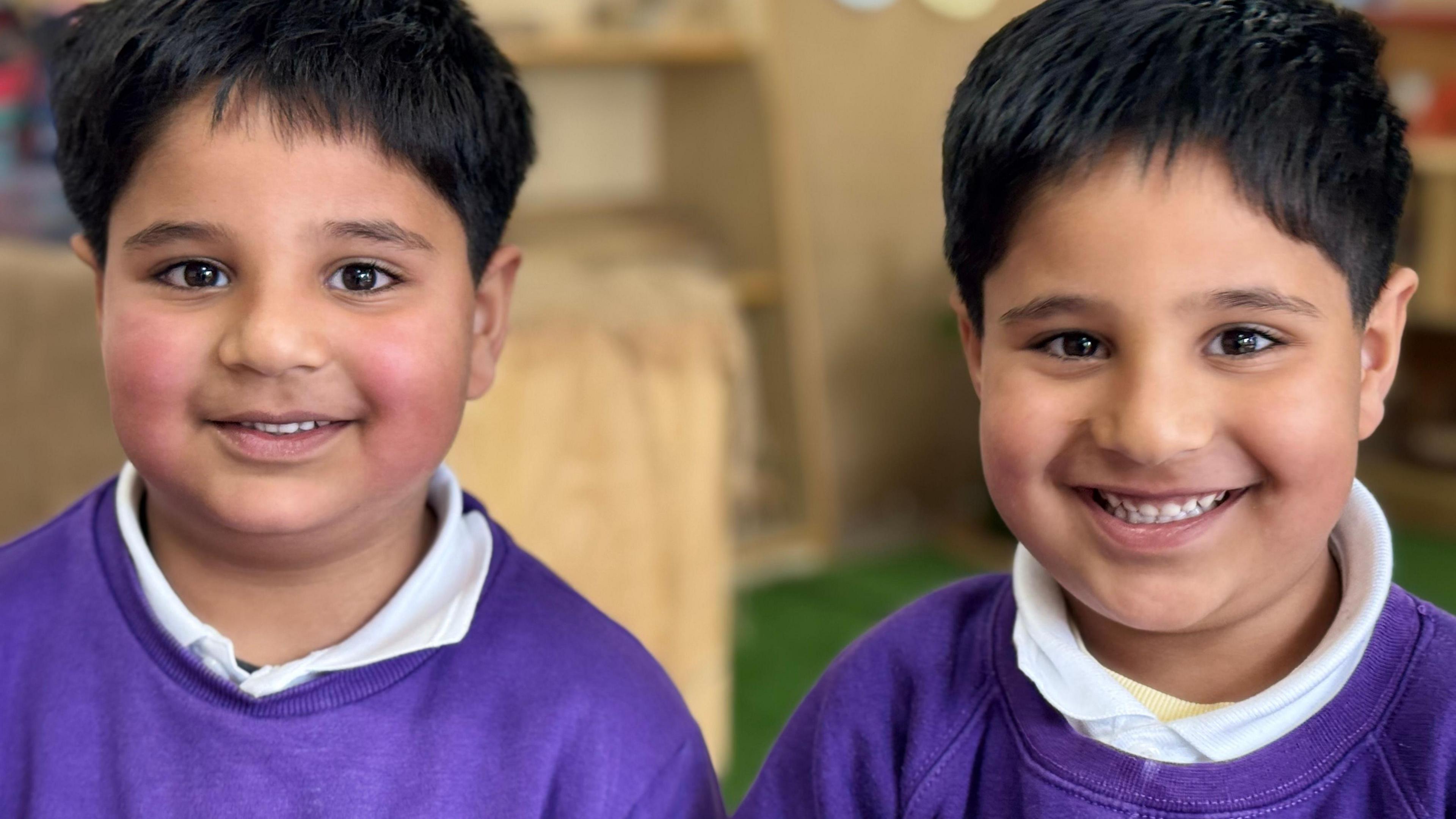 Two boys smile into the camera with short black hair. They both wear purple tops with white collars.