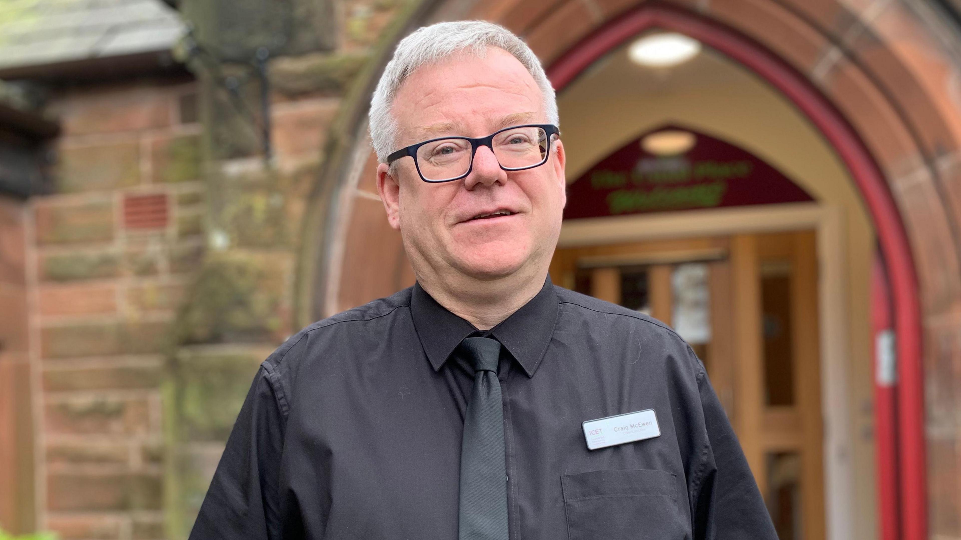 A grey haired man in glasses with a black shirt and tie stands in front of an arched sandstone doorway