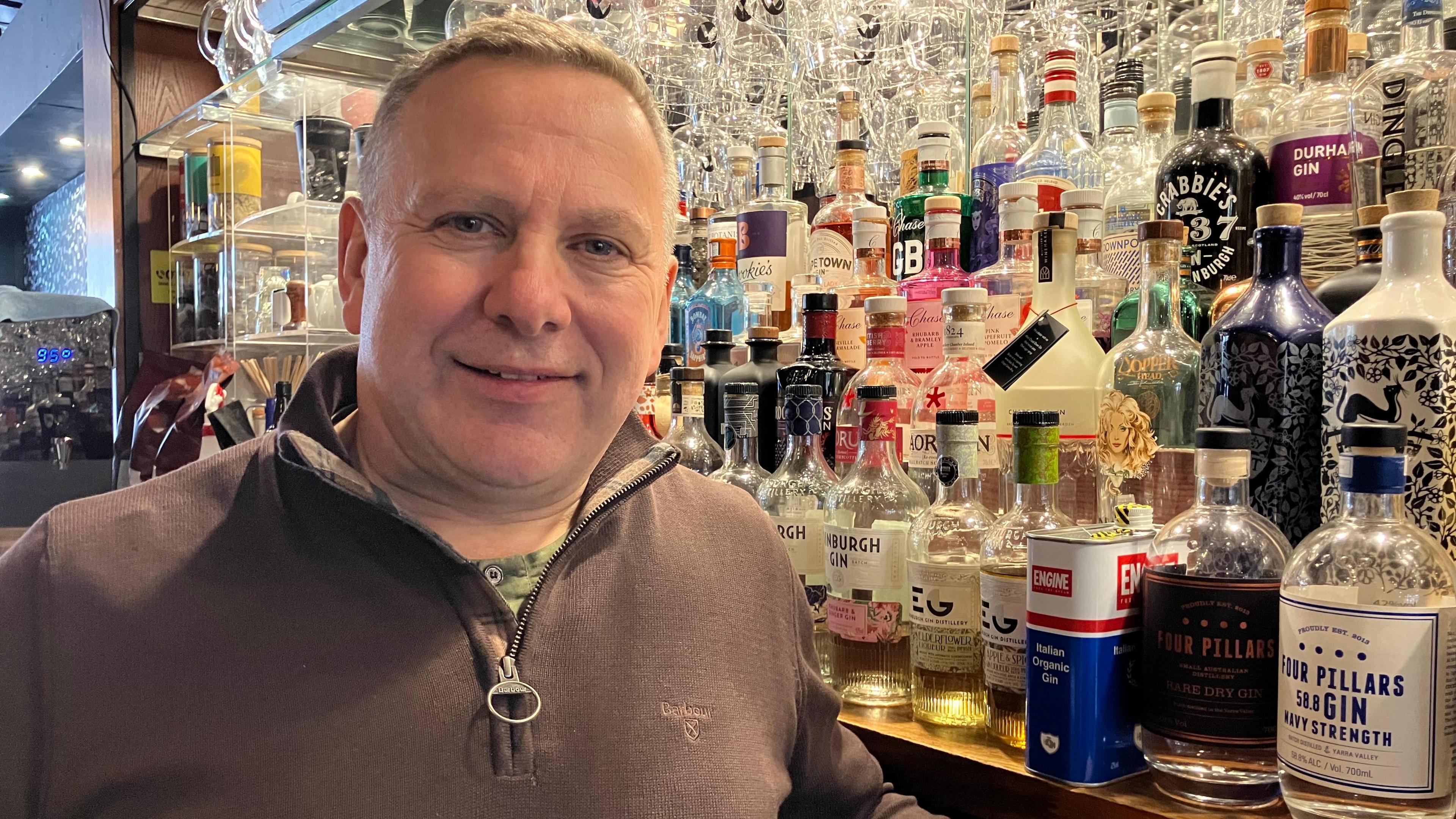 Mark Wrigley, with grey hair and wearing a grey/brown fleece, is standing behind the bar smiling. Behind him is a wide selection of gin bottles.