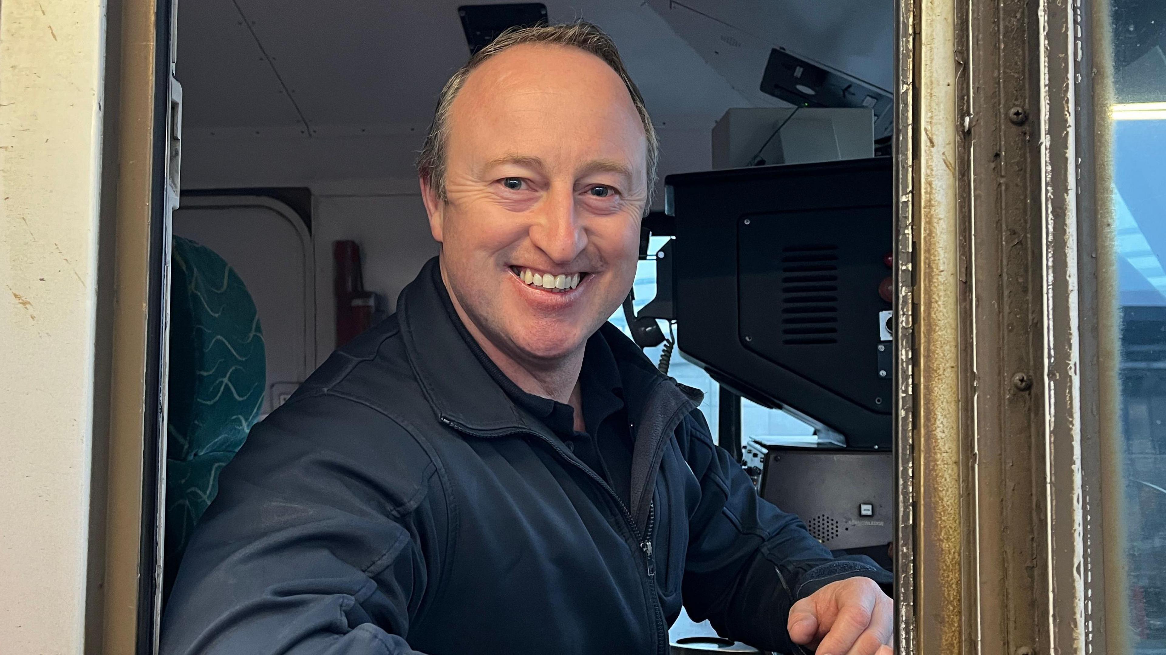 A train driver smiles into the camera. He's sitting on the driver's seat on a train. He's wearing a navy uniform.