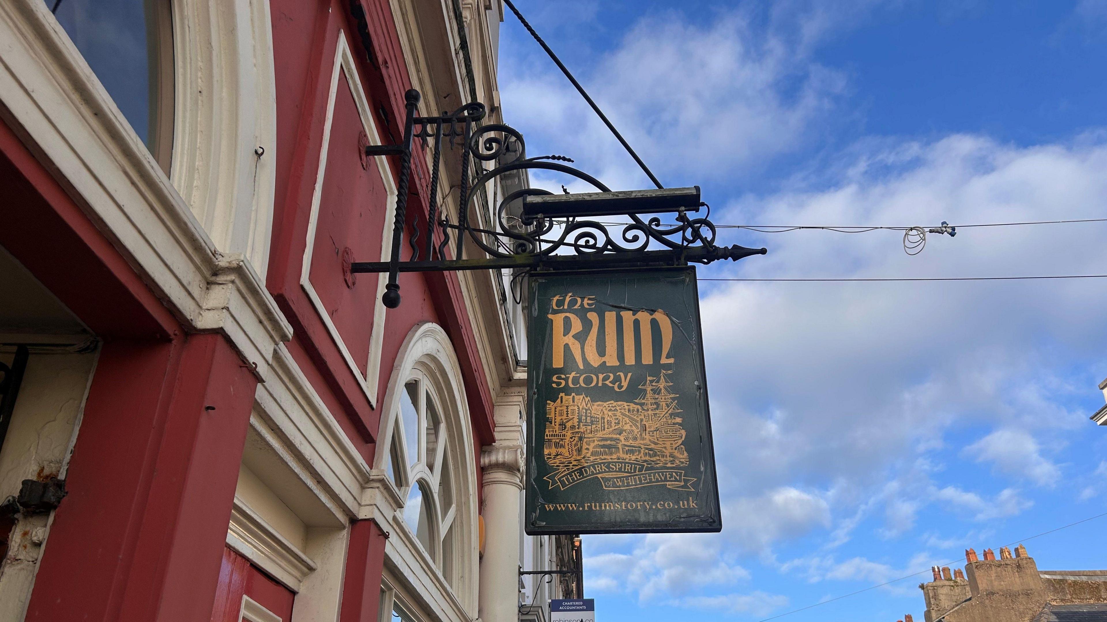 Sign saying "The Rum Story" attached to red brick building. The sign is green, the lettering orange and the image is often several ships. 