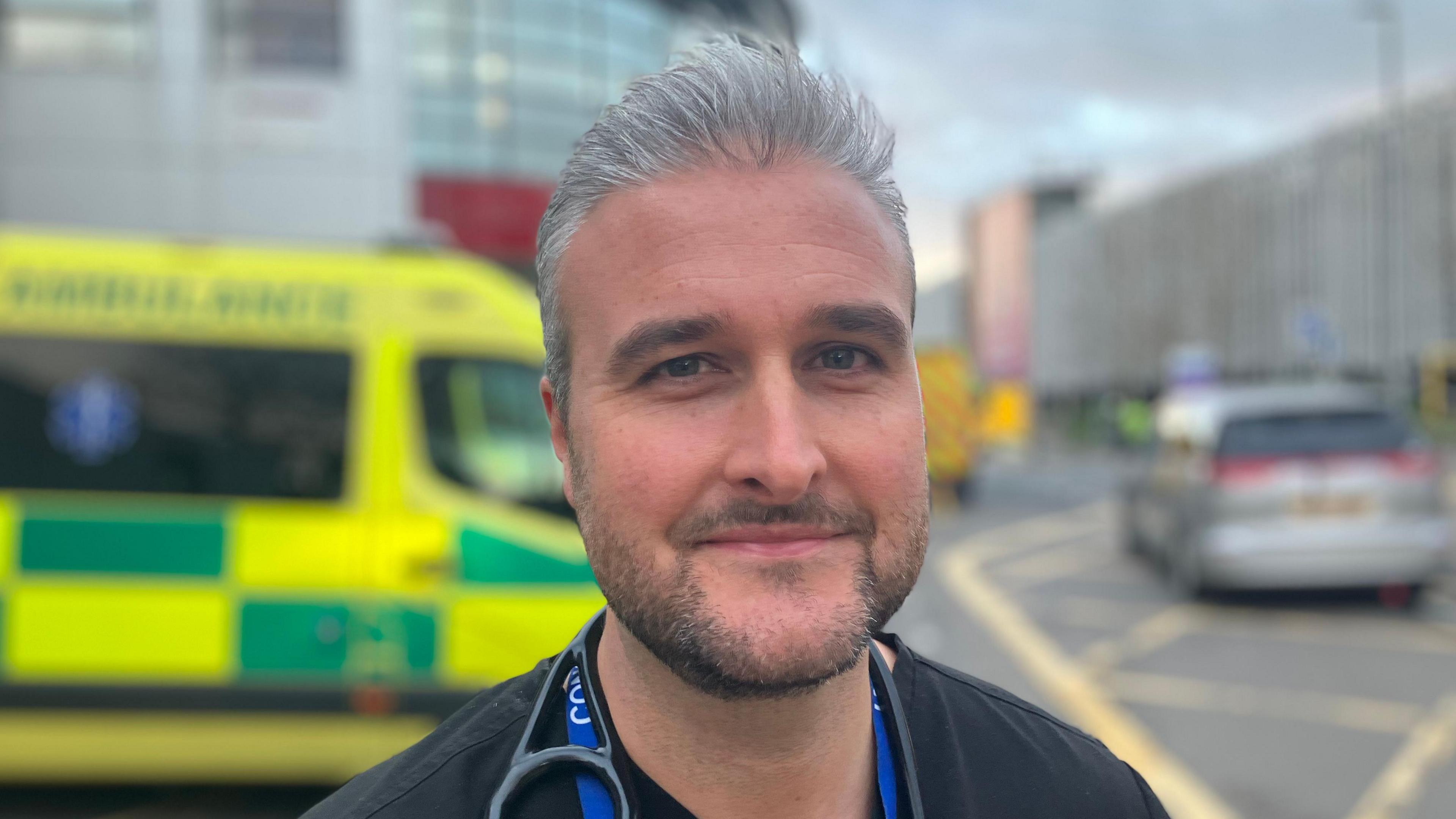 A man with grey hair and a short beard stood outside. There is an ambulance and other cars behind him. He is wearing a dark top.