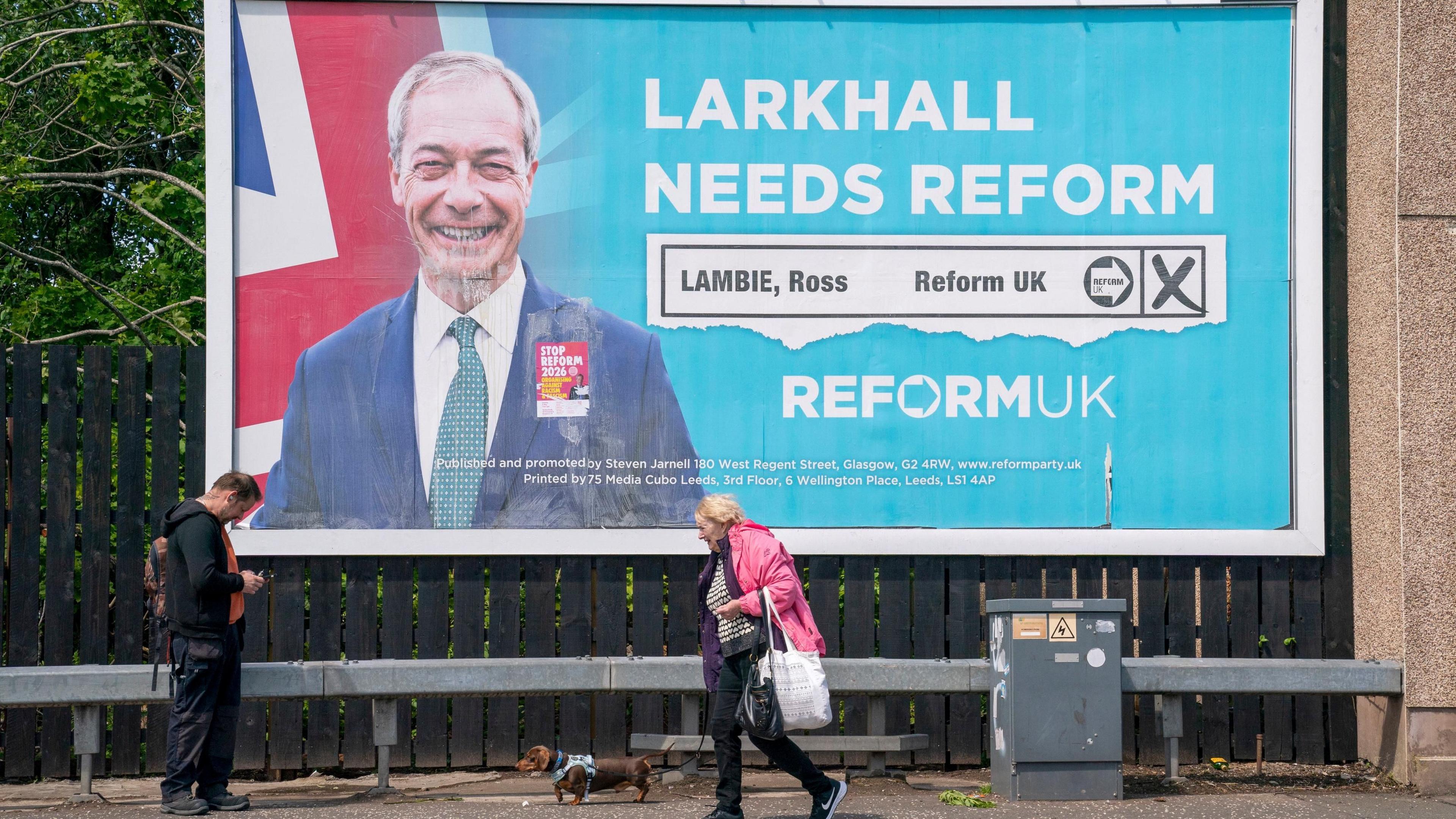 Reform UK election billboard poster in Larkhall, featuring a picture of Nigel Farage and the words Larkhall Needs Reform. A woman in a pink jacket is walking a dog from right to left and a man in black clothes is looking at his phone on the left of the picture.