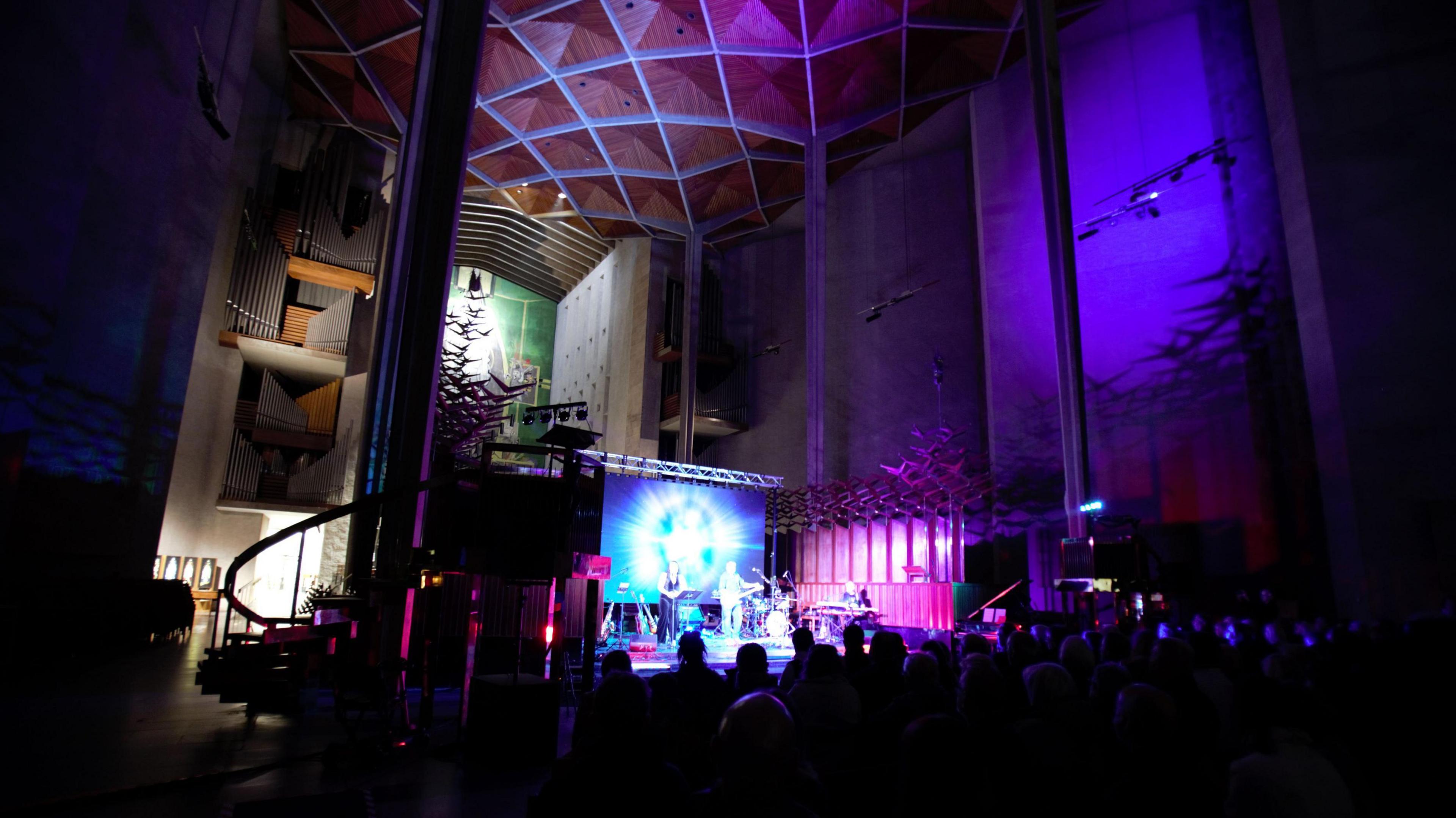 A band can be seen inside the cathedral with the audience in front of them. In the background is the organ and above the band are high ceilings and ti the side is a staircase. Behind the band is a screen which is lit-up in blue and white light. 