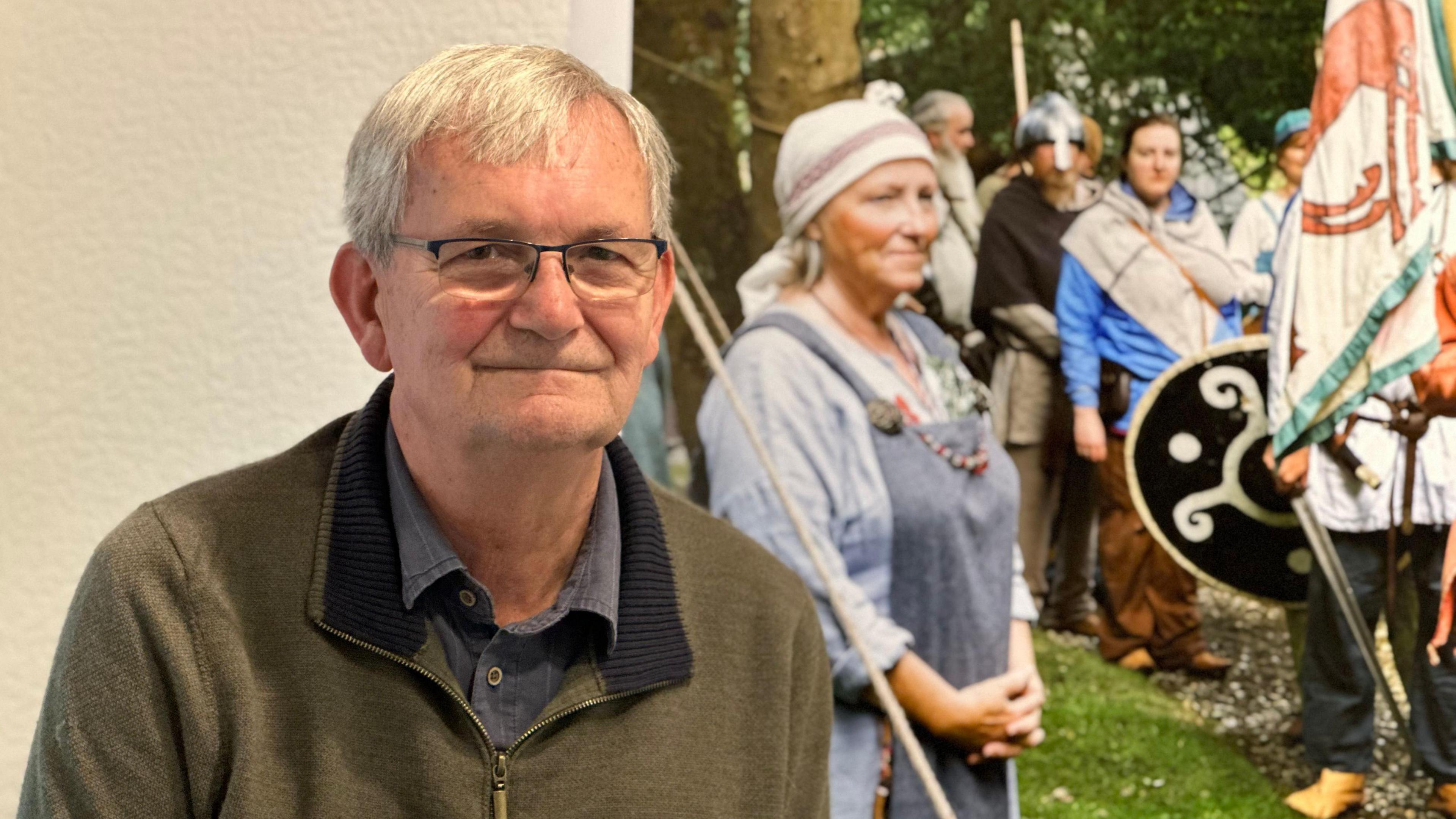 An elderly man wearing glasses with grey hair with a friendly smile, wearing a dark green jumper, with people dressed like vikings in the background.