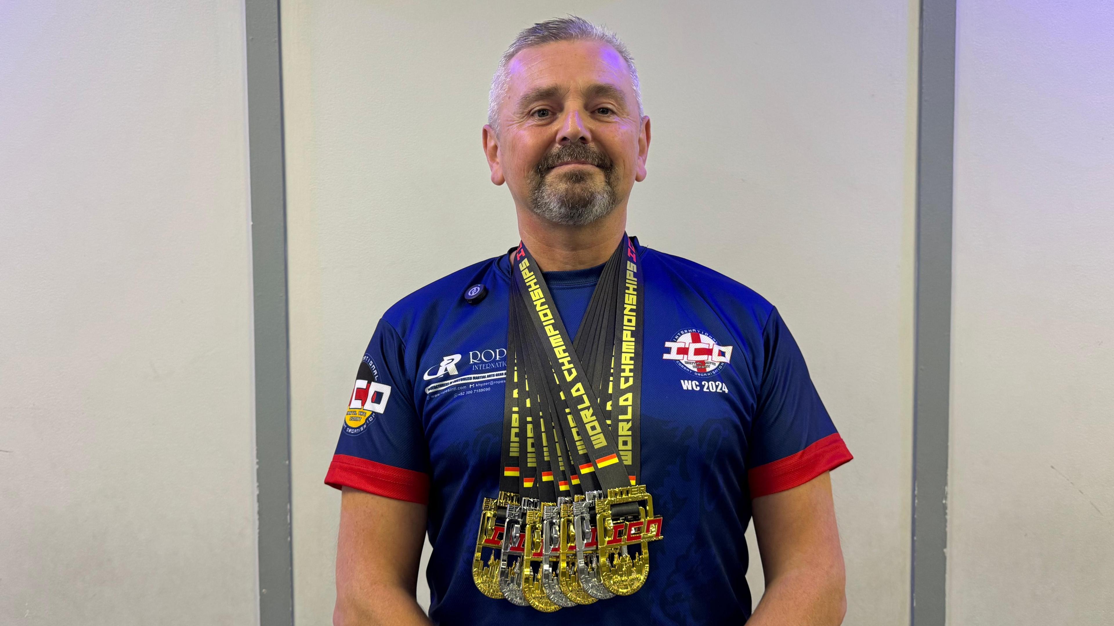 A man with short grey hair and beard wearing a blue and red T shirt and several medals around his neck.