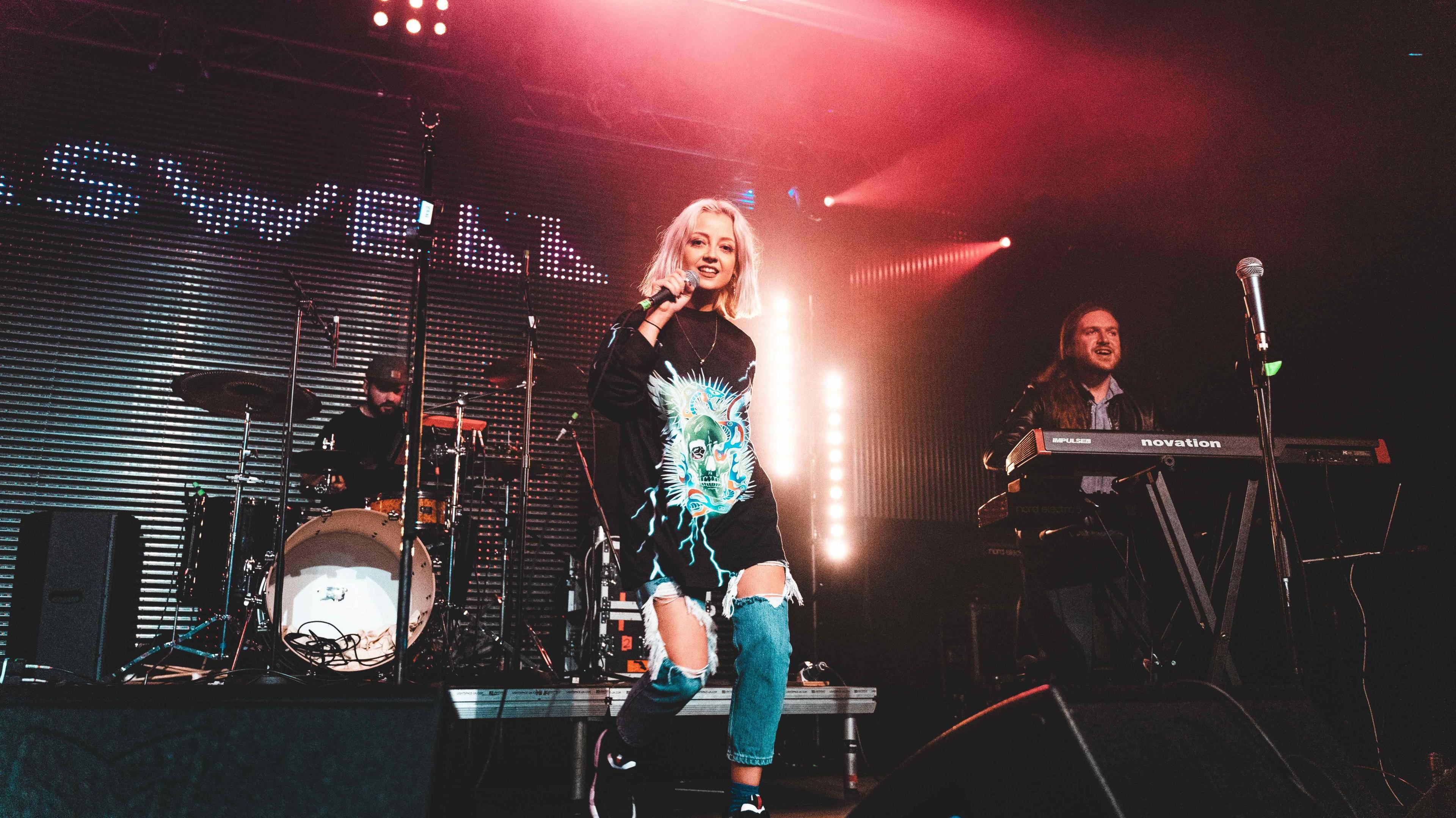 Caswell on a stage with ripped jeans, a keyboard and drum kit behind