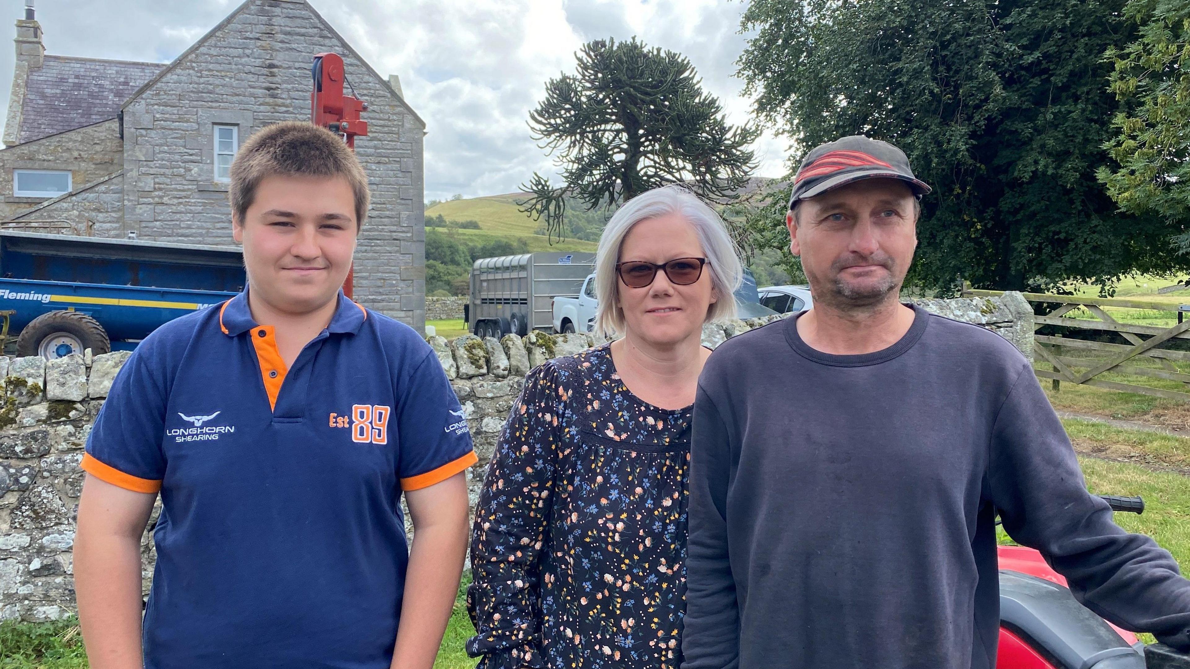 Shona and Scott Anderson stand next to their son T-jay Anderson. He is about 16 and wearing a blue top with short dark hair. She has ash blonde hair and has a blue flowery top on, Scott is wearing a cap, has a grey top and is about 40. Behind there is a stone wall and a Victorian house