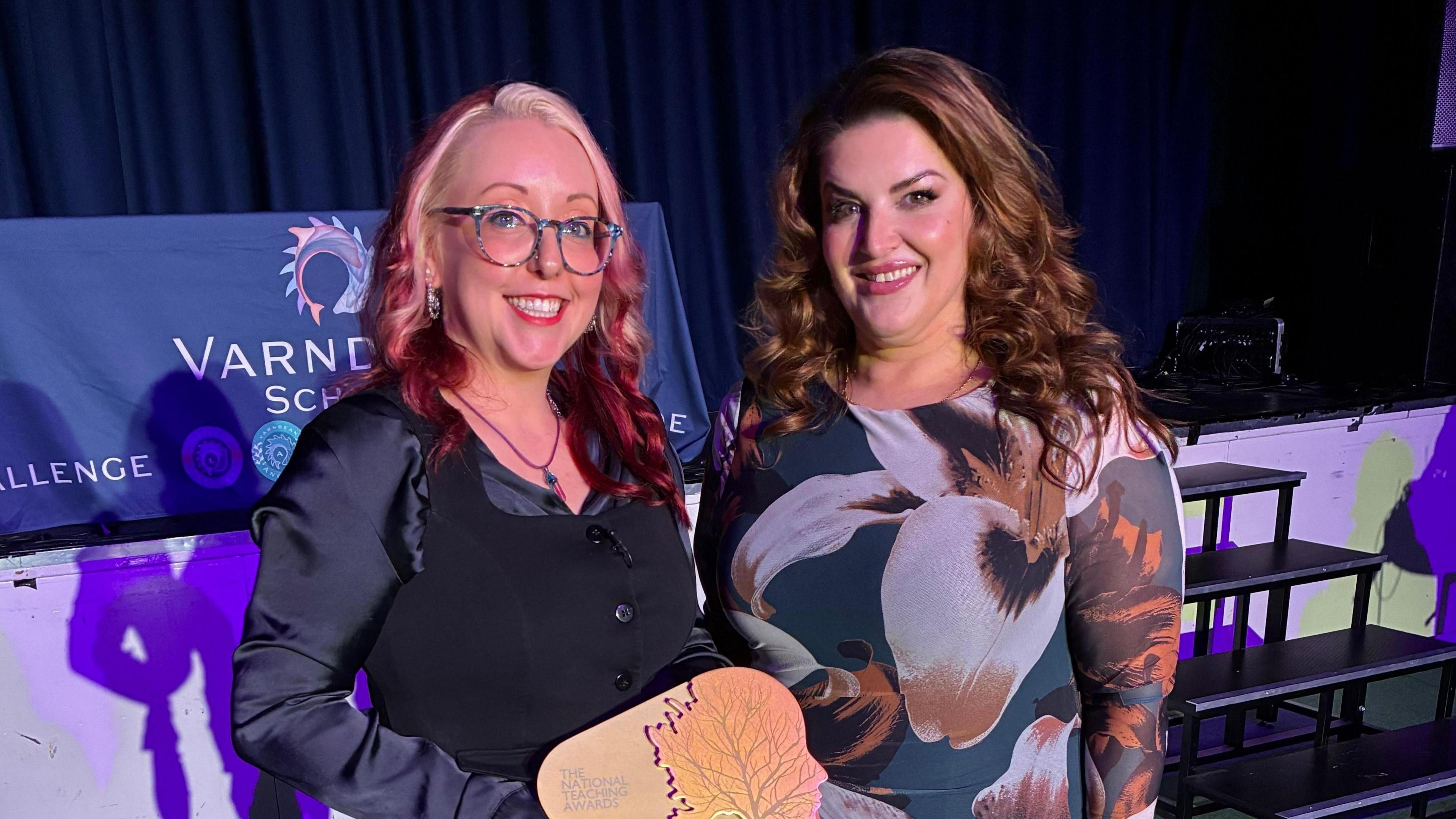 Jodie Prenger stands to the right. She has long brown curled hair and is smiling at the camera. Jolie Bartley stands on the left. She has red hair with white face framing pieces. They are curled. She is wearing blue round glasses and holding a trophy, smiling at the camera. 