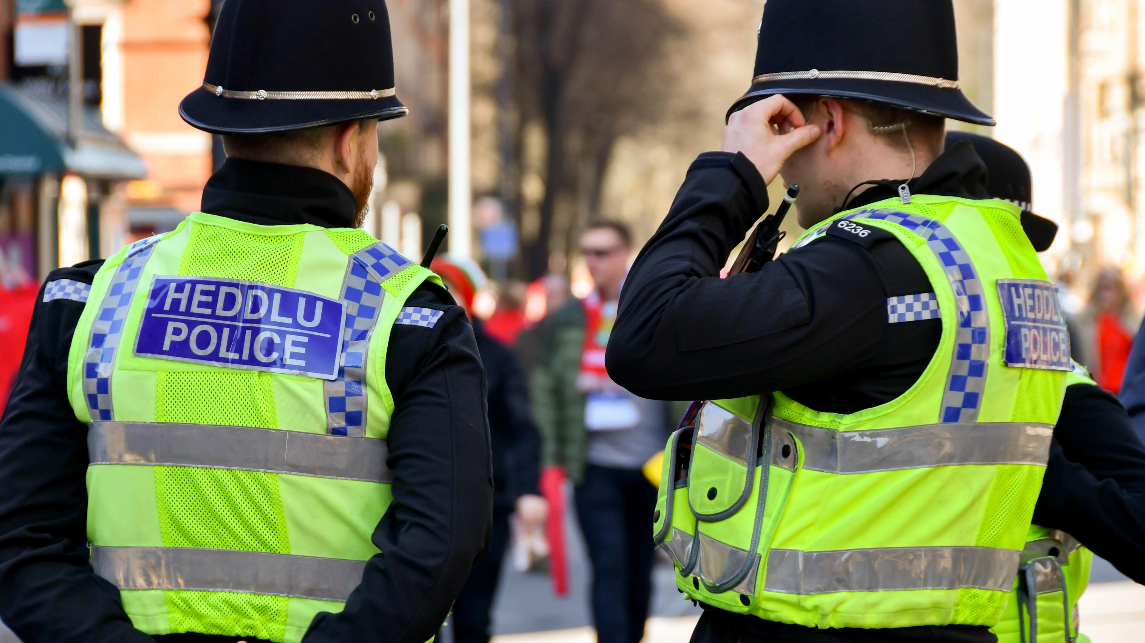 Two police officers stand facing away from the camera. They are wearing fluorescent vests with the words heddlu and police on the back and are wearing black helmets. 
