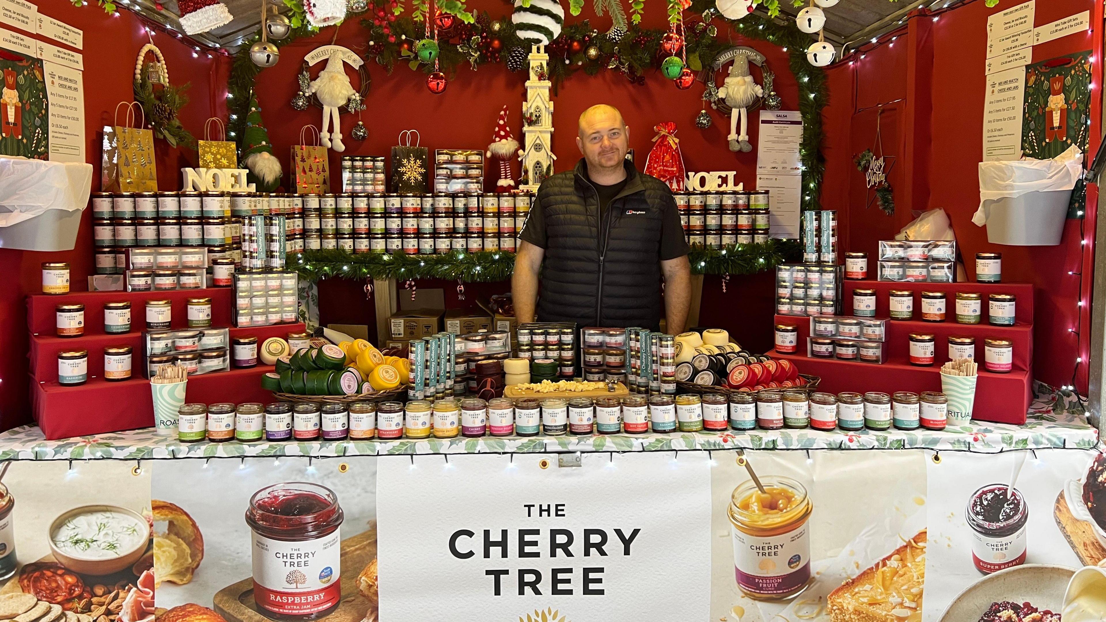 A bald man, wearing a black t-shirt and black body warmer stands in the middle of a cheese and preserves stall. He is smiling and is surrounded by Christmas decorations and jars of preserves and blocks of cheese. 