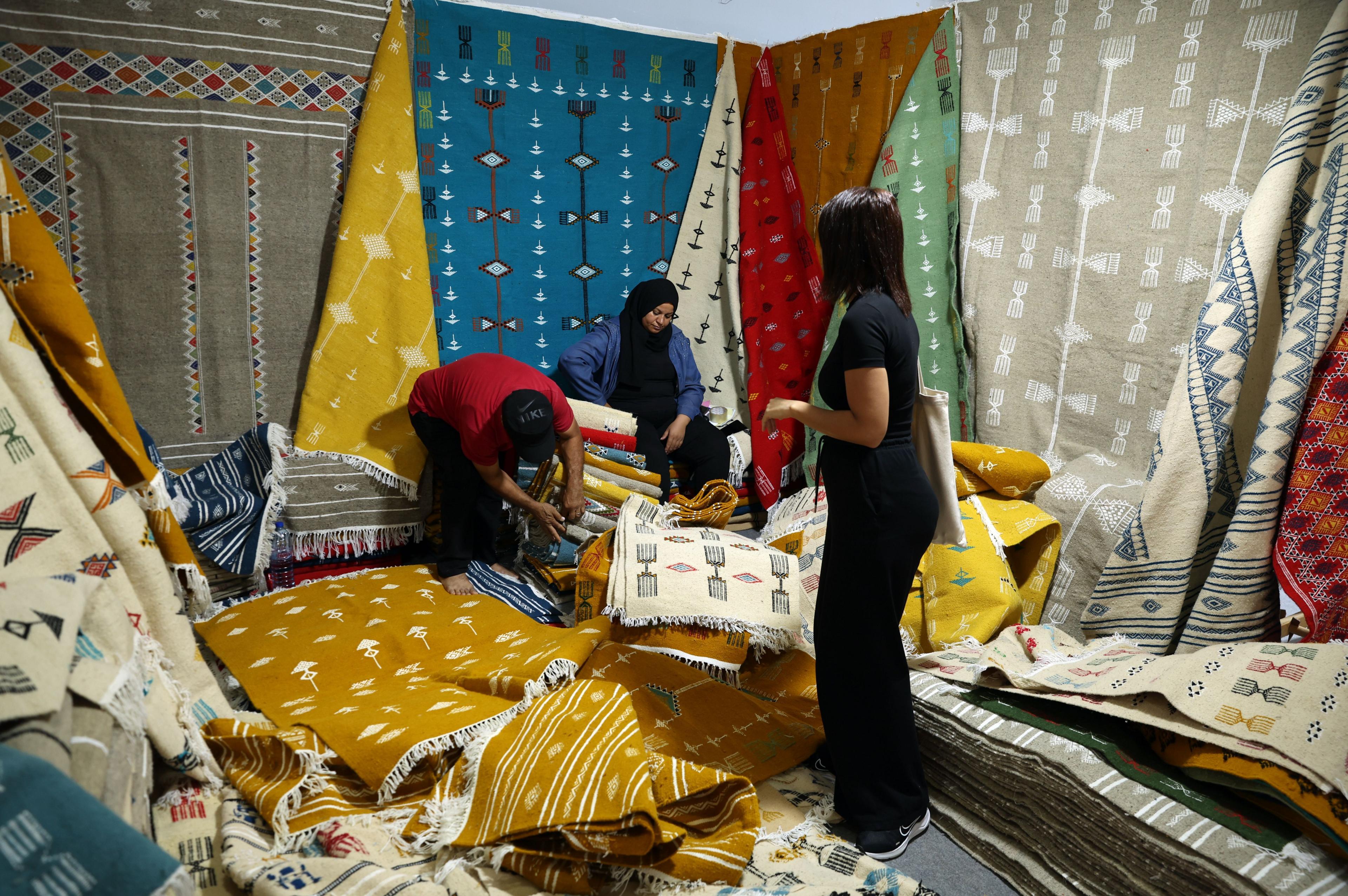 Women look a various brightly coloured rugs for sale in a large exhibition booth.