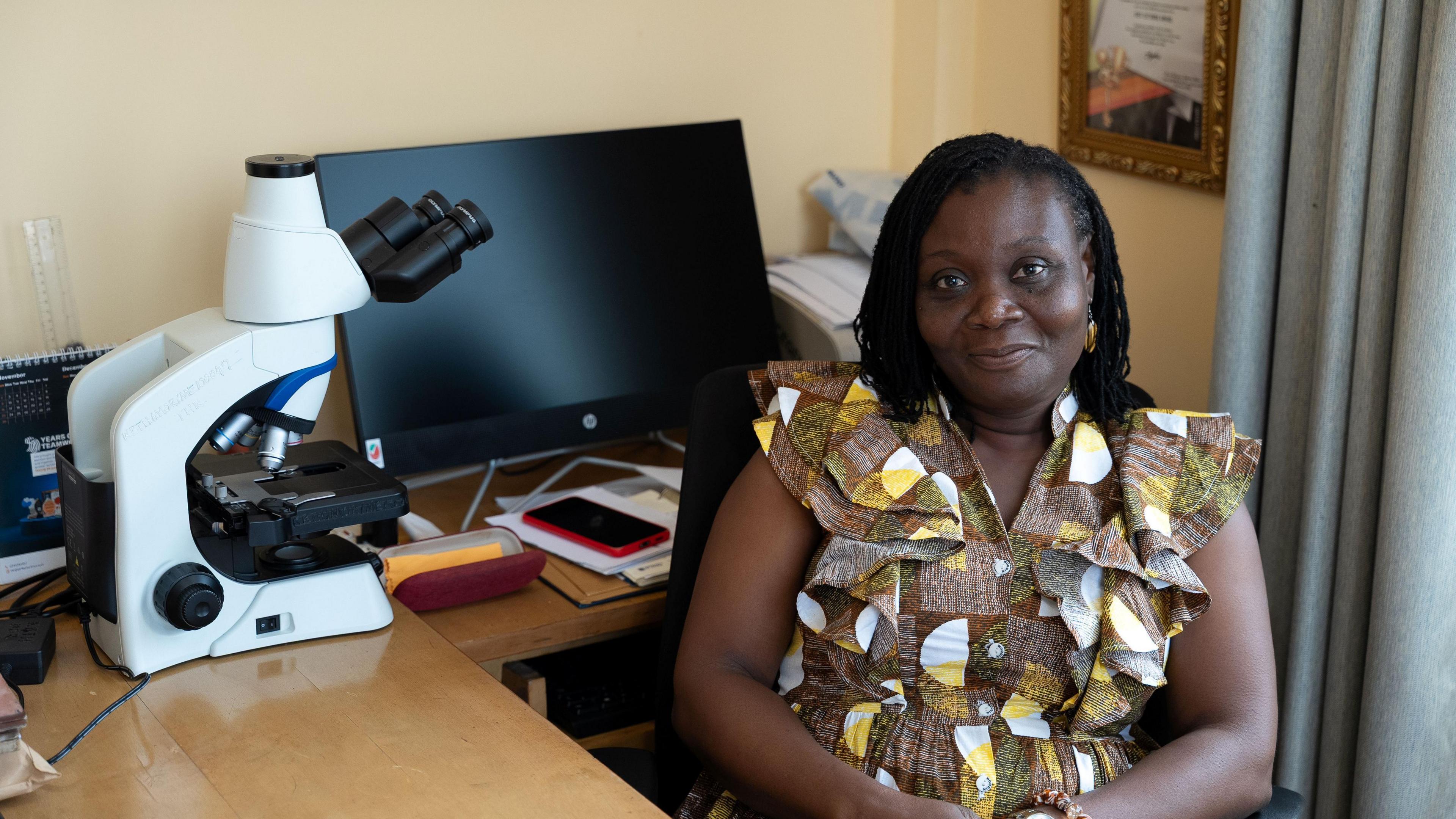 A woman sitting at a desk with a microscope and a computer monitor in a room.