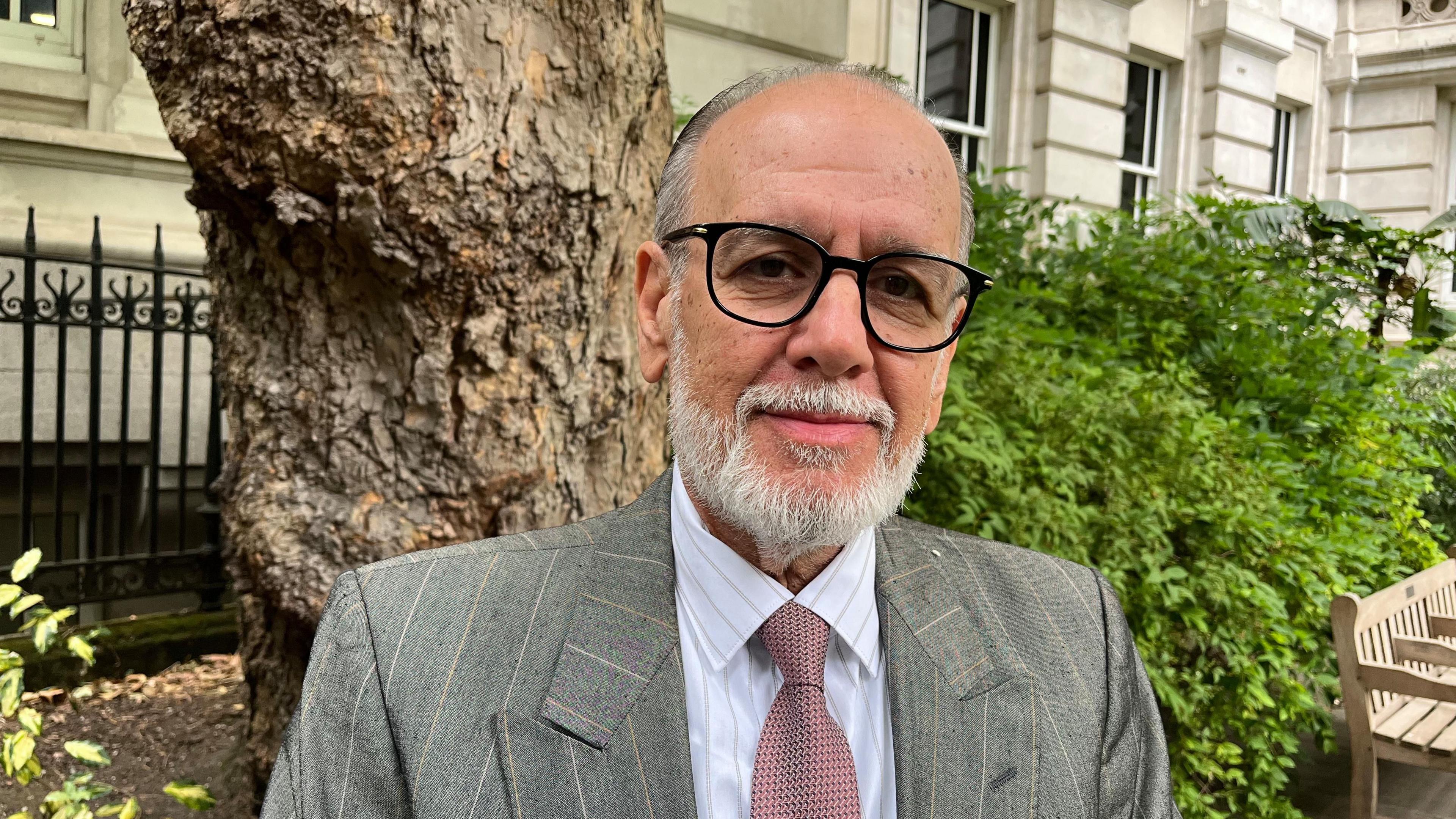George Psaradakis in a mauve tie, grey suit and black glasses standing in a park with a tree trunk behind him.