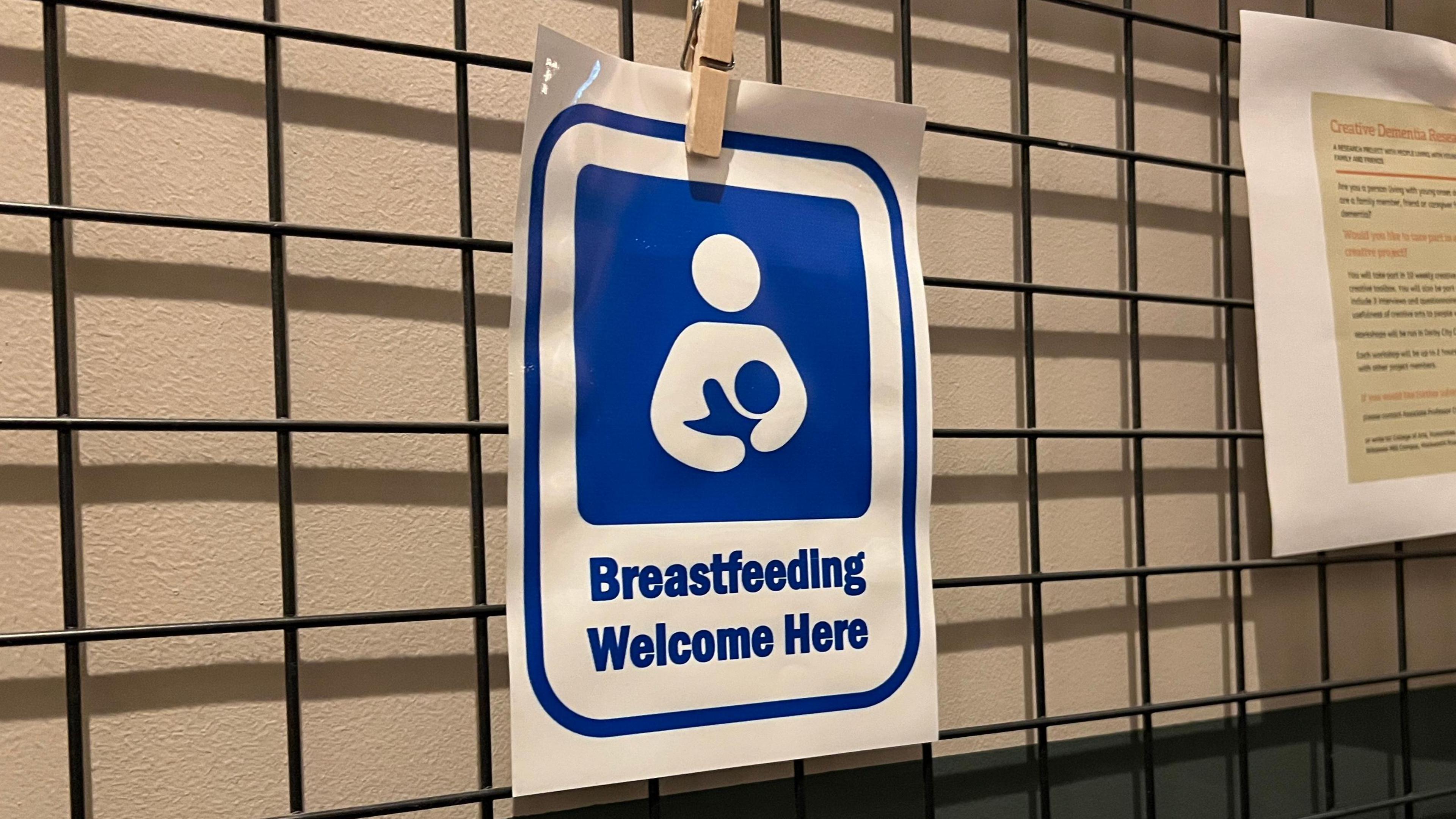 Blue and white sign saying "Breastfeeding welcome here" is pegged on rails in front of a white wall