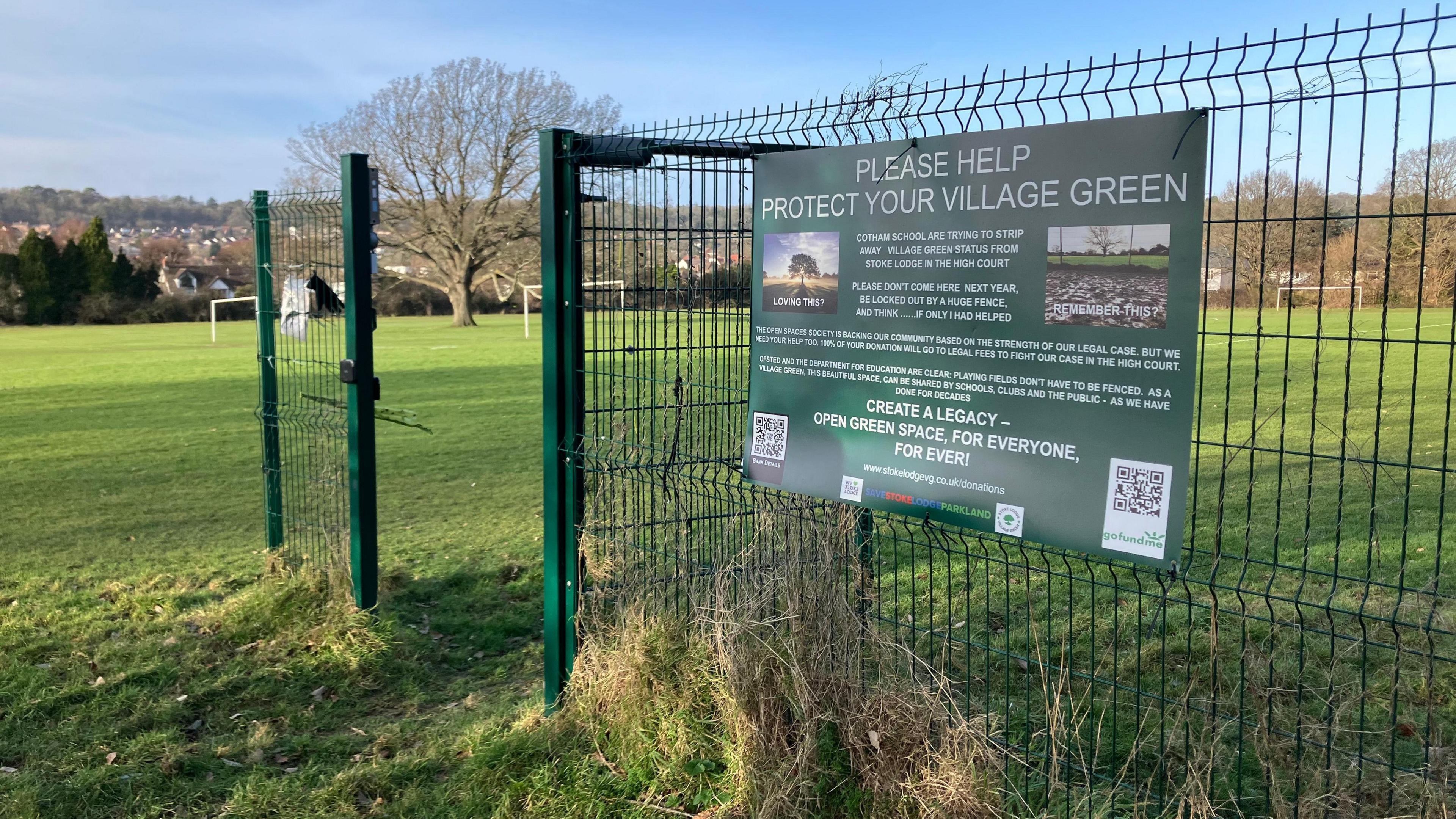 A green fence is seen along a green field. There are goal posts in the distance. A sign on the fence says Please help protect your village green.