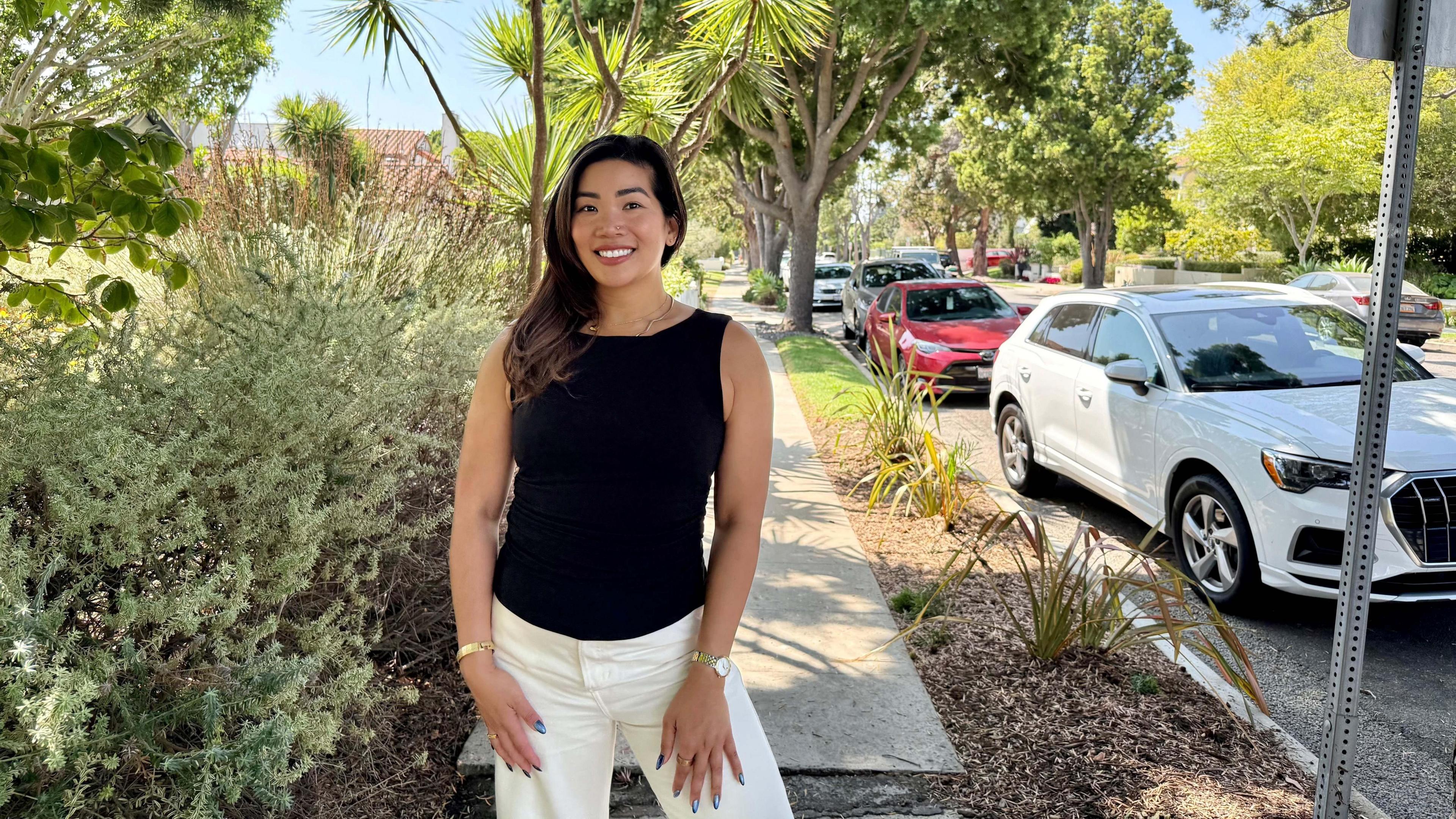 A woman wearing a black shirt and white pants poses on the sidewalk of a tree-lined residential street, next to a parked white car.
