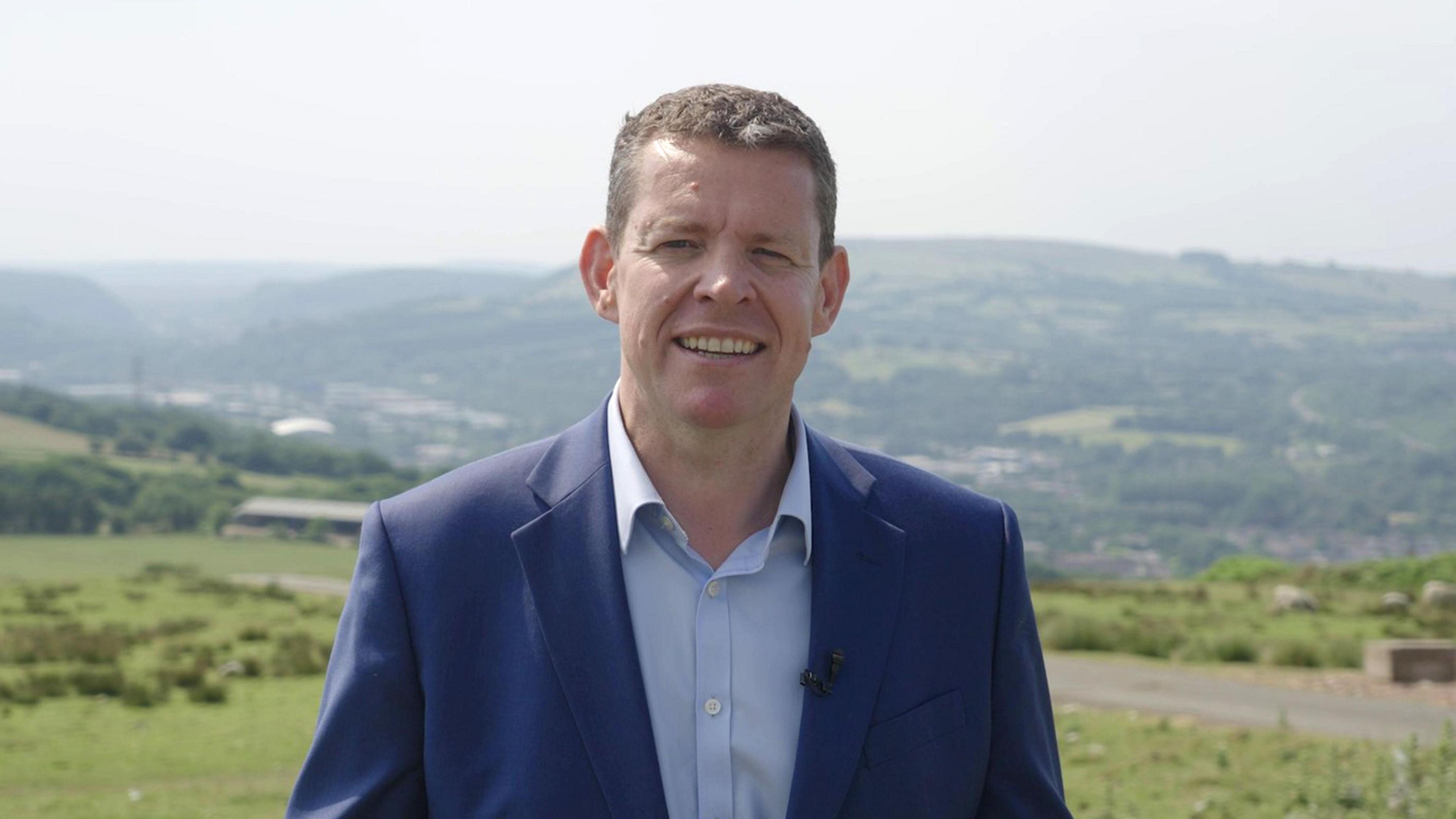 Rhun ap Iorwerth yn gwisgo siwt glas tywyll a chrys glas golau, gyda bryniau a chaeau gwyrdd yn y cefndir.