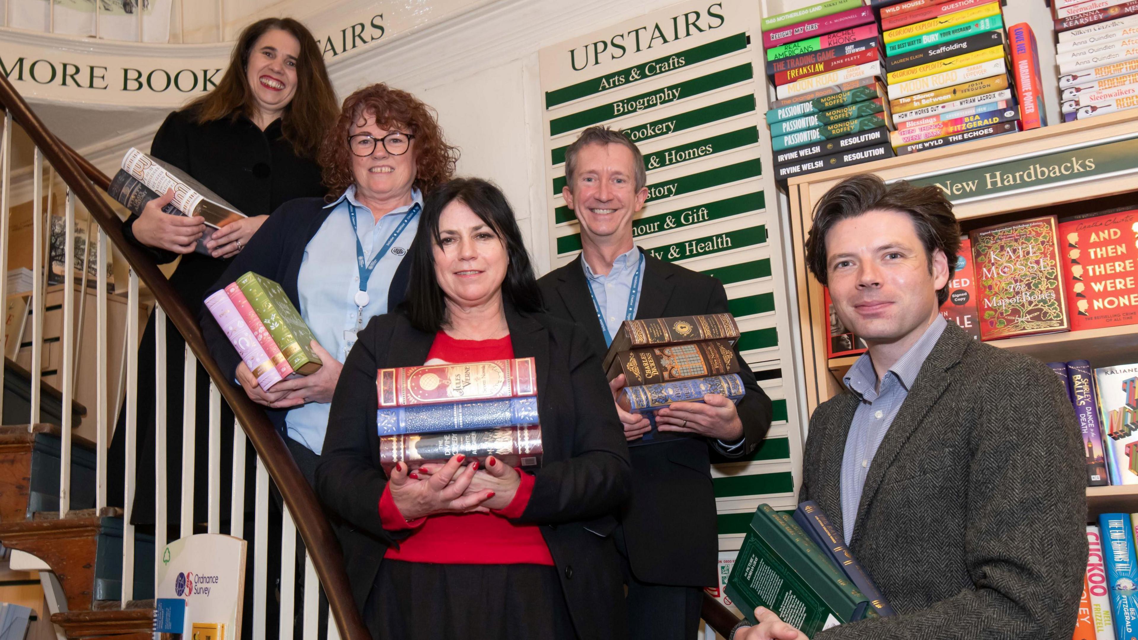 Three women and two men hold piles of books on the staircase inside a book shop.