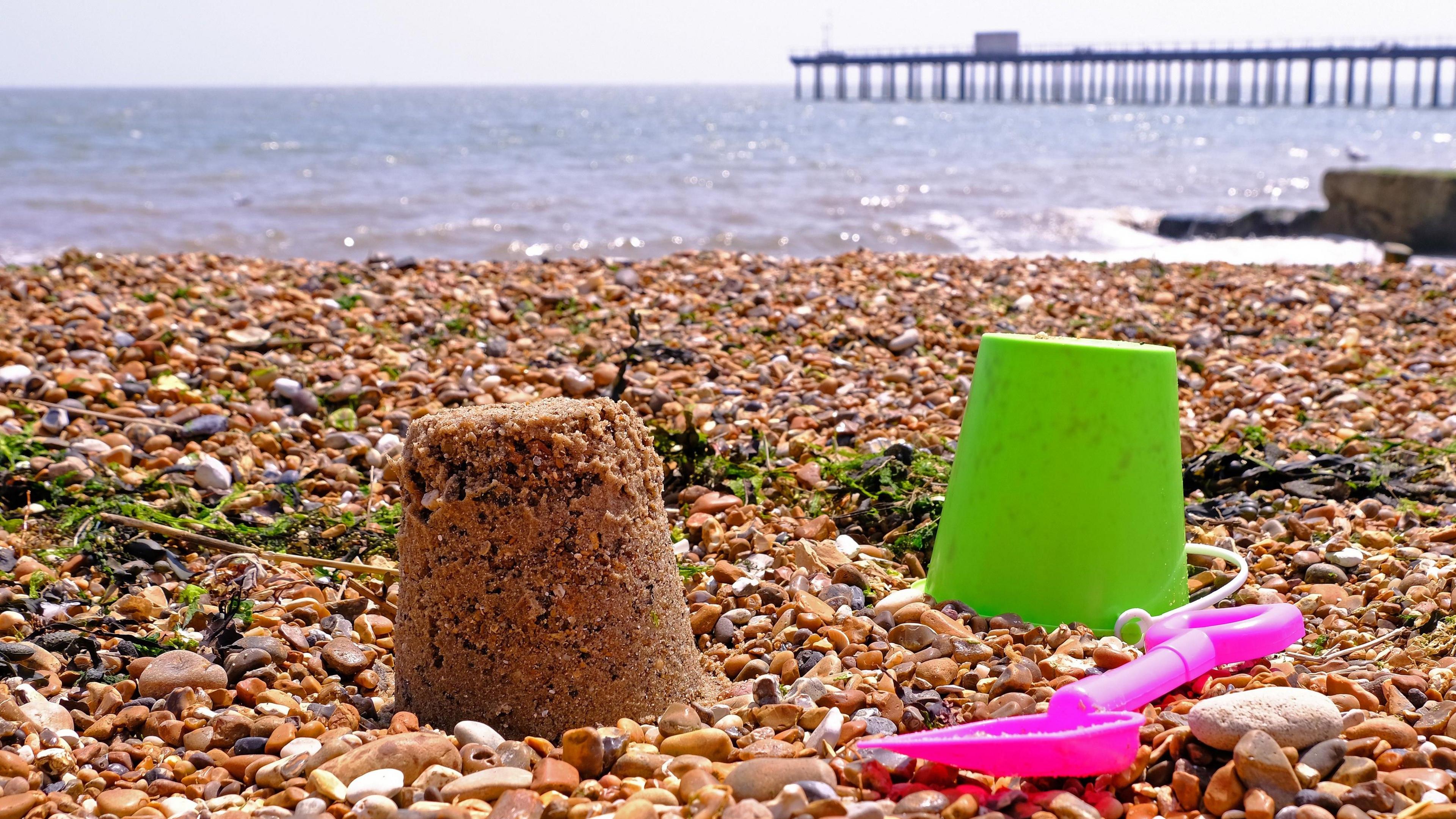 A sandcastle rests on a shingle beach. A green bucket and pink space rest next to it. A pier can be seen in the background leading into the sea.