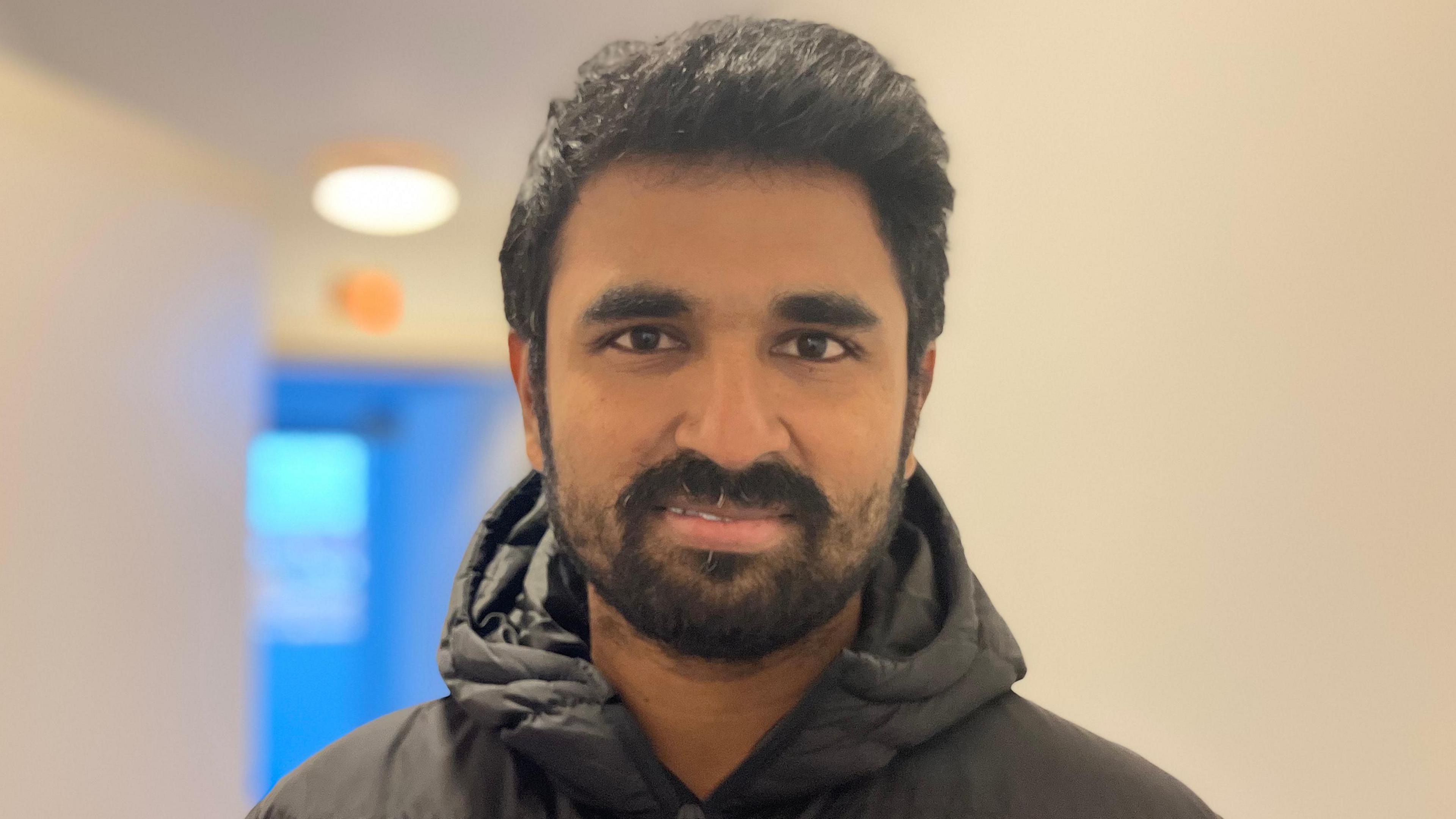 Harsha Kunku wearing a black coat and has a beard. The background is blurry showing the corridor of his home in the Mosaic Apartment block.