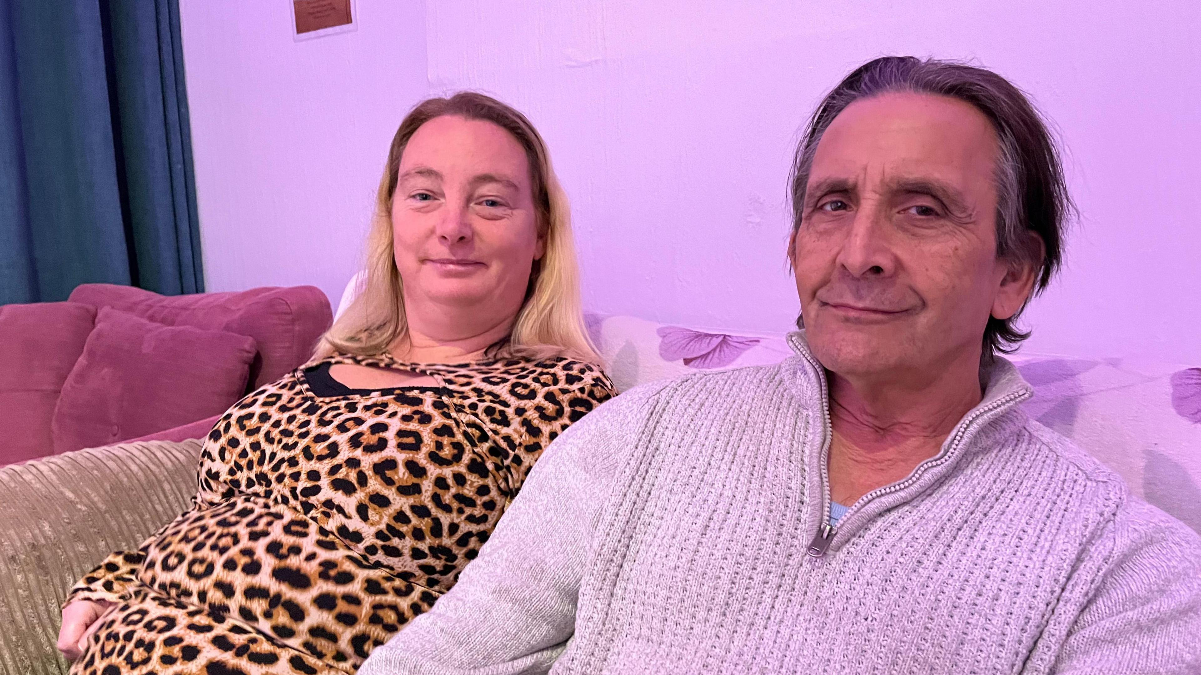 Phoebe's mum, Tamzin, and dad. The mum has a leopard print top on and has blonde hair. The dad has a grey jumper with grey hair.