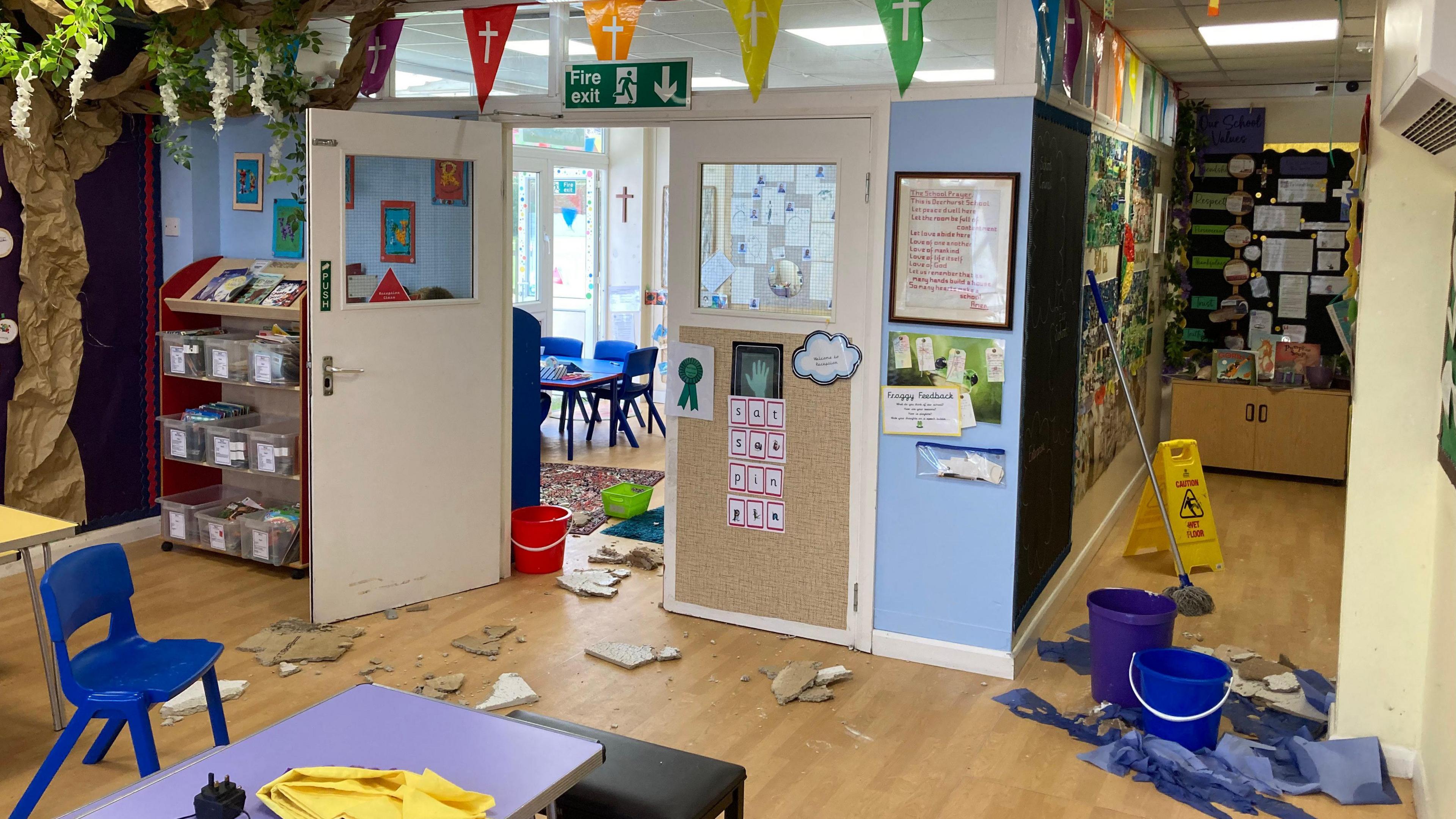 A school with piece of broken ceiling on the floor. There is a mop and bucket on the right hand side with some wet blue towels and a wet floor sign