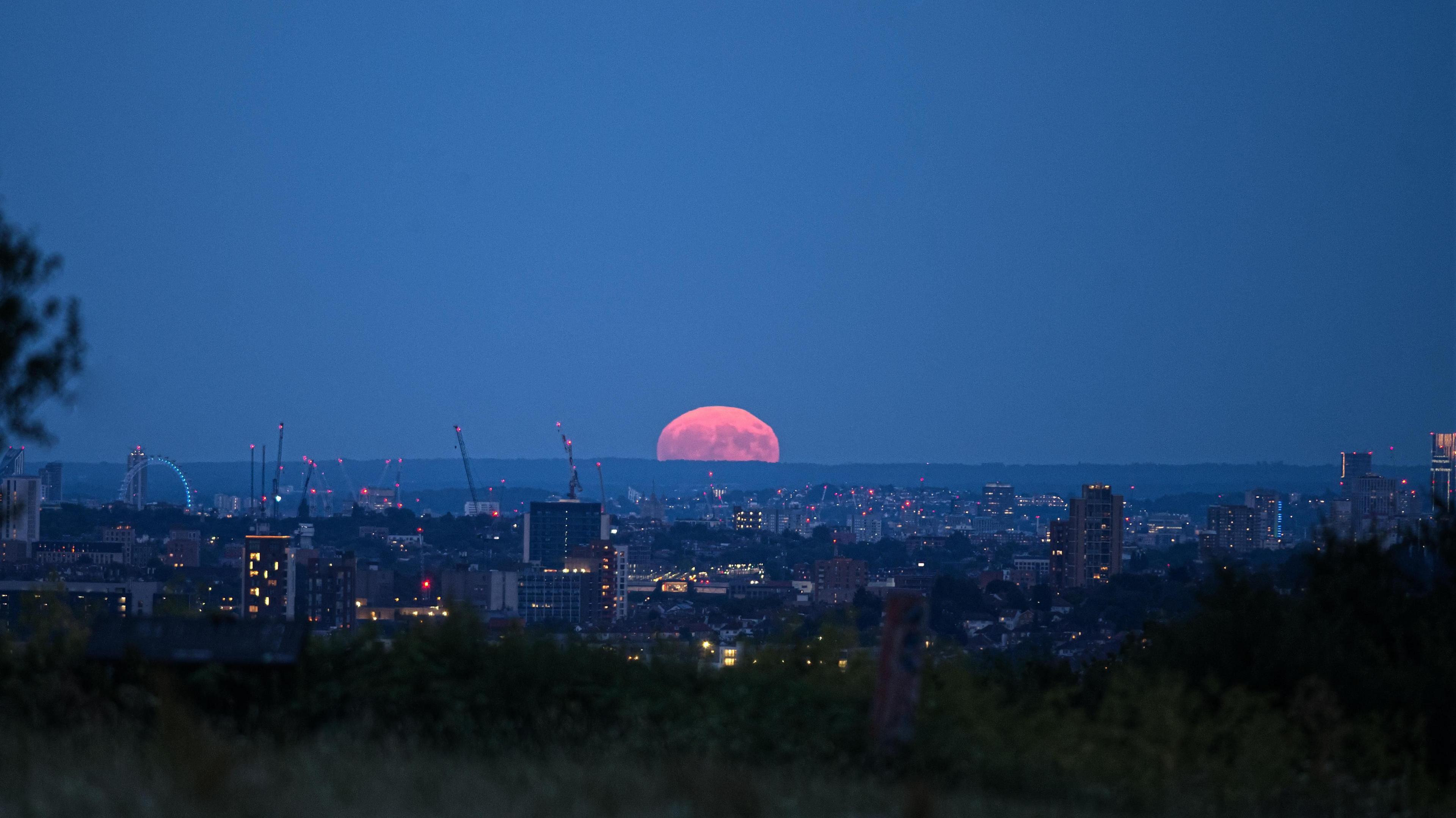 An orange-hued Strawberry Moon sits on the horizon with a city in the foreground